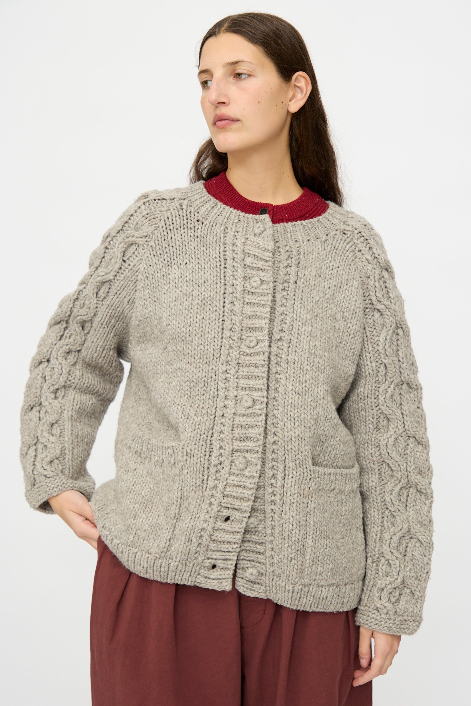 A woman wears the nest Robe Peruvian Wool Cable Sleeve Knit Cardigan in Beige, featuring a chunky cable-knit design, oversized fit, and front pockets, over a red top and loose burgundy pants against a plain white background.