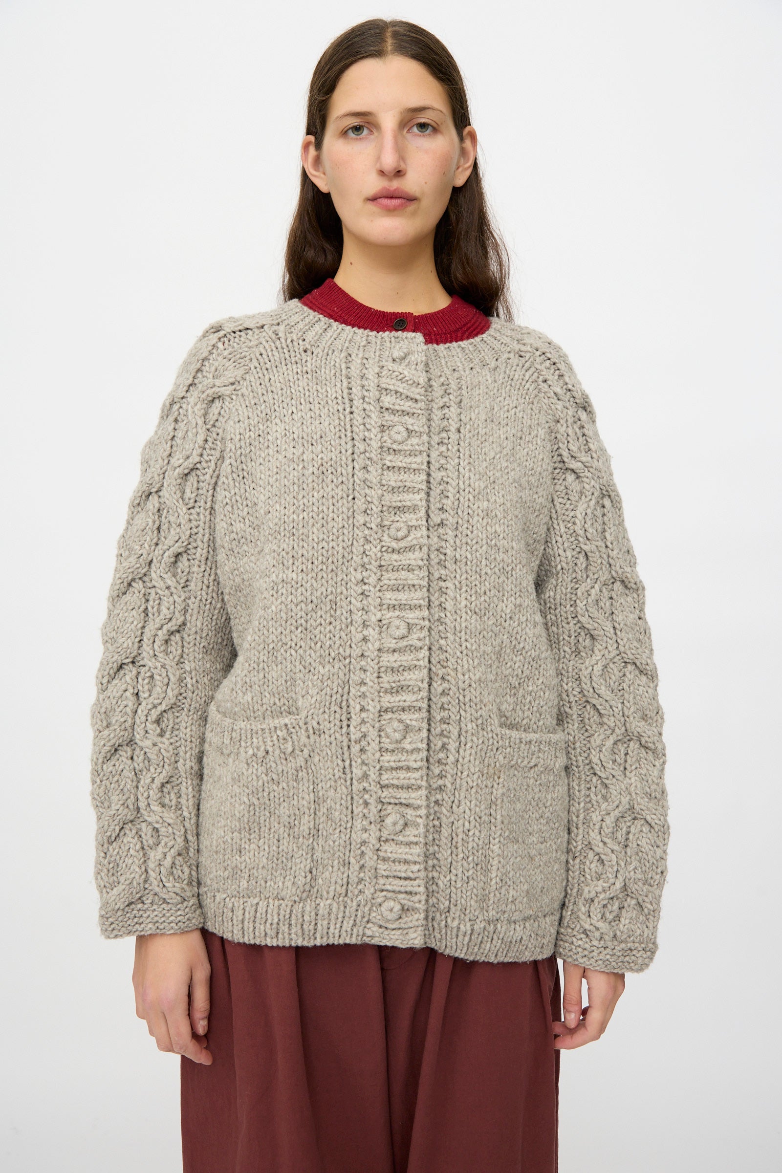 A woman faces forward wearing the nest Robe Peruvian Wool Cable Sleeve Knit Cardigan in Beige over a red top and maroon skirt, posed against a plain white background.