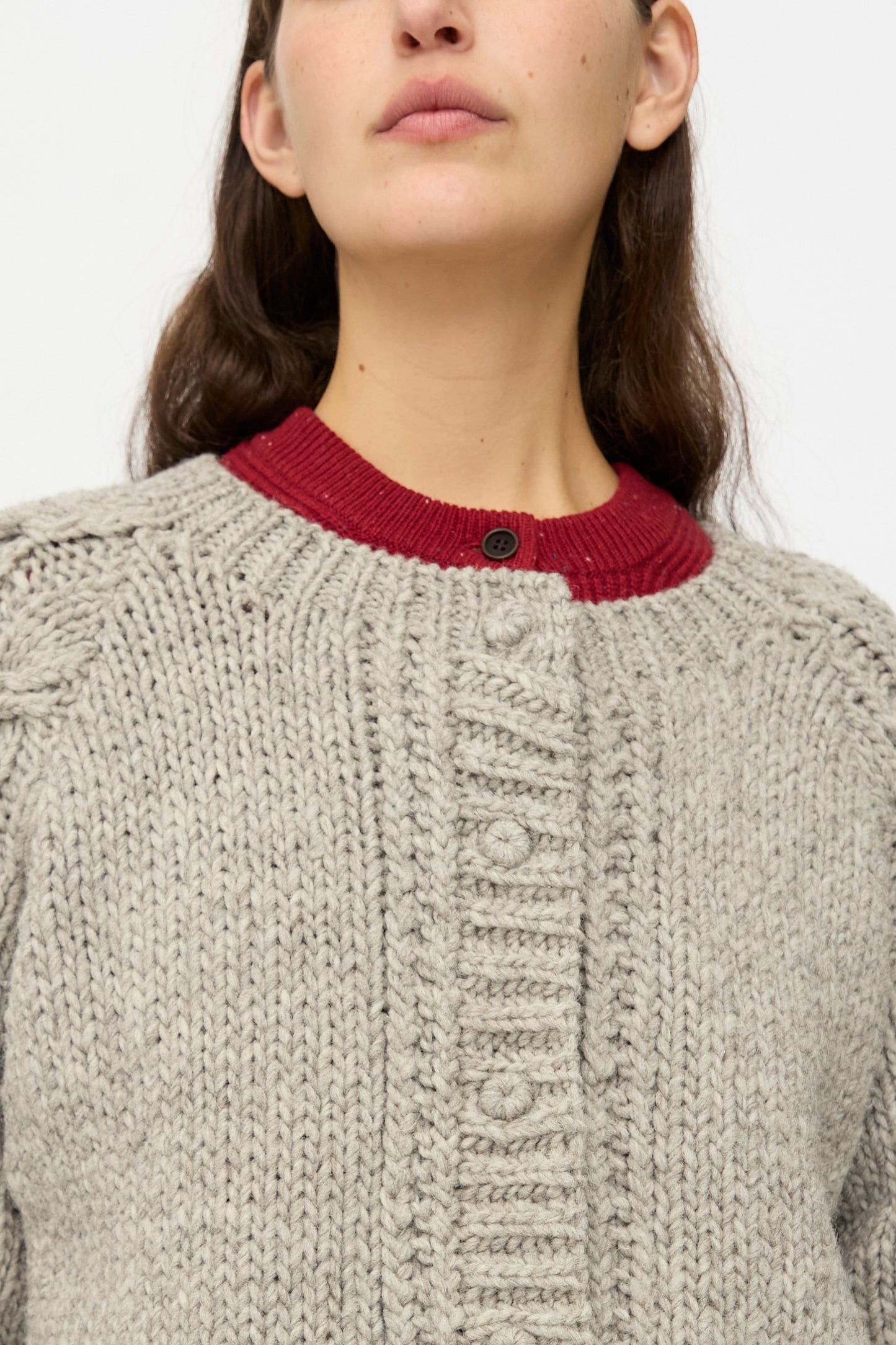 A woman wears the nest Robe Peruvian Wool Cable Sleeve Knit Cardigan in Beige over a red crew neck sweater, photographed from the shoulders up against a plain background.