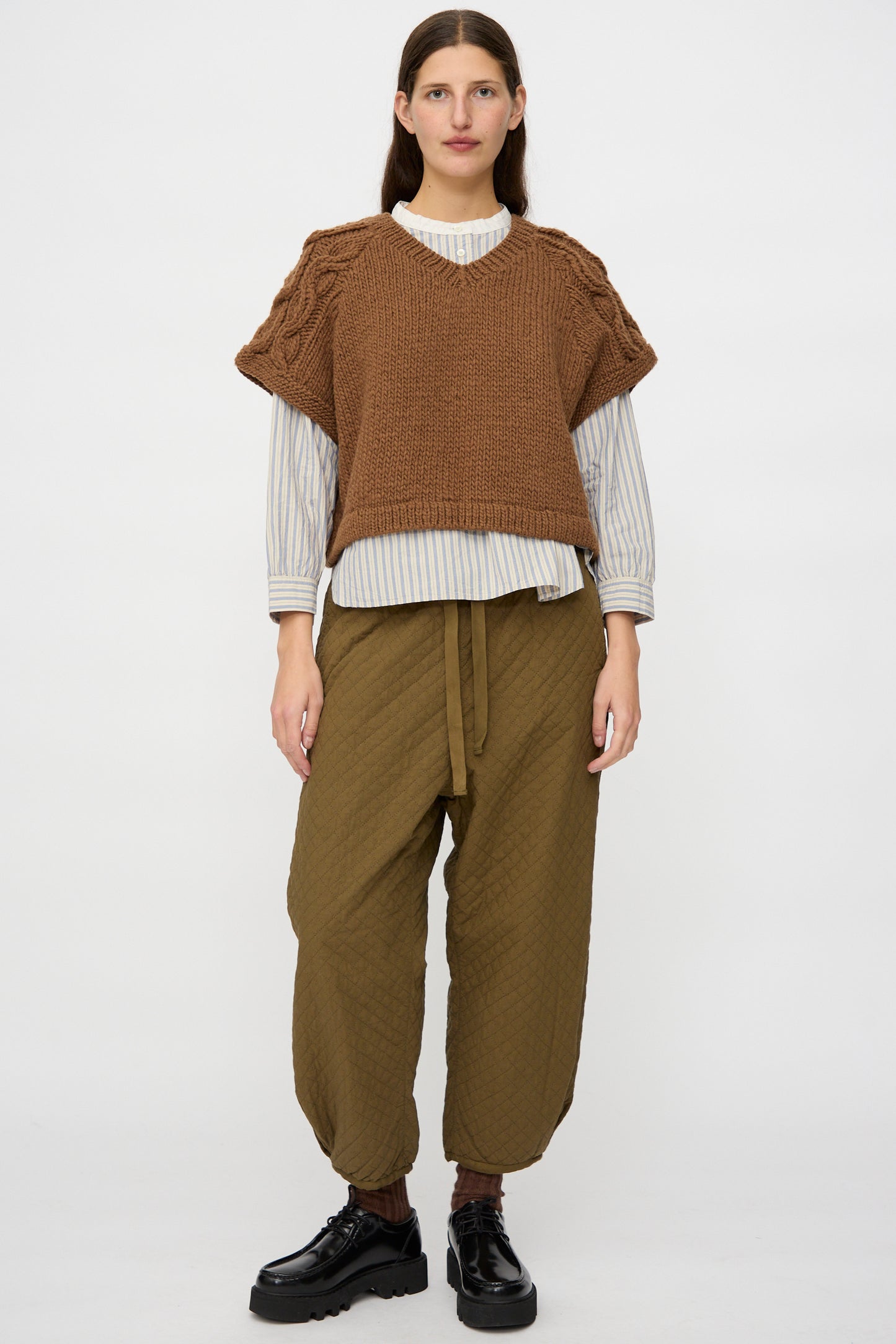 A person stands facing forward in a nest Robe Peruvian Wool Raglan Cable Knit Vest in Camel over a striped shirt, loose olive tie-waist pants, and black shoes against a plain white background.