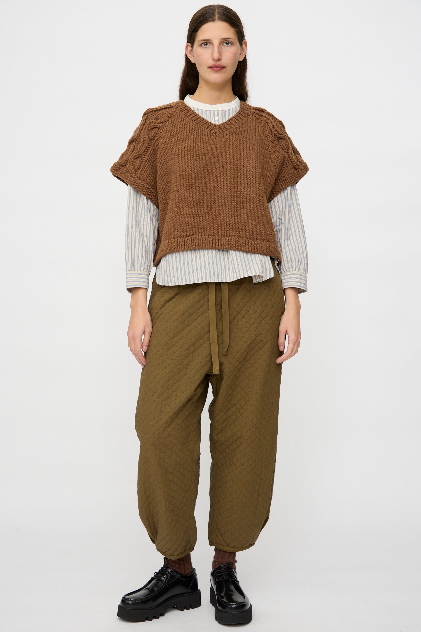 A person stands facing forward in a nest Robe Peruvian Wool Raglan Cable Knit Vest in Camel over a striped shirt, loose olive tie-waist pants, and black shoes against a plain white background.