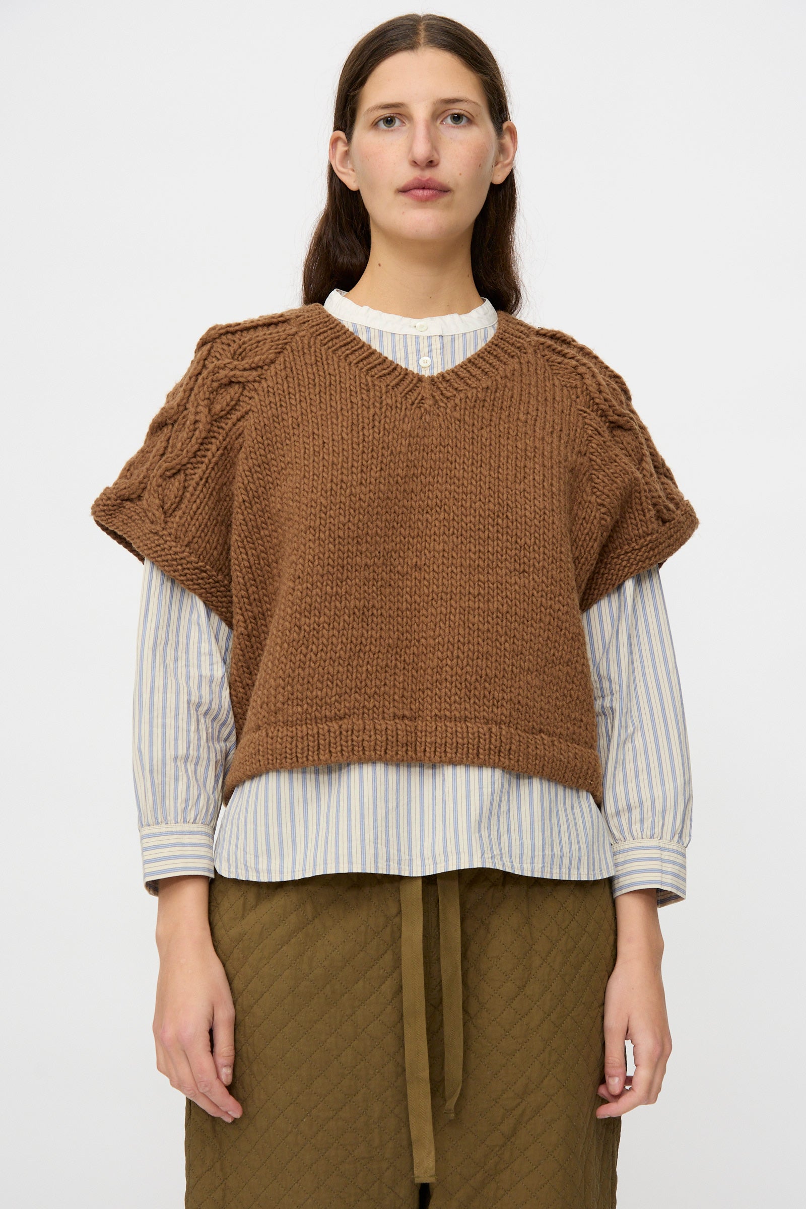 A model wears the nest Robe Peruvian Wool Raglan Cable Knit Vest in Camel over a striped shirt with an olive quilted skirt, standing against a plain white background.