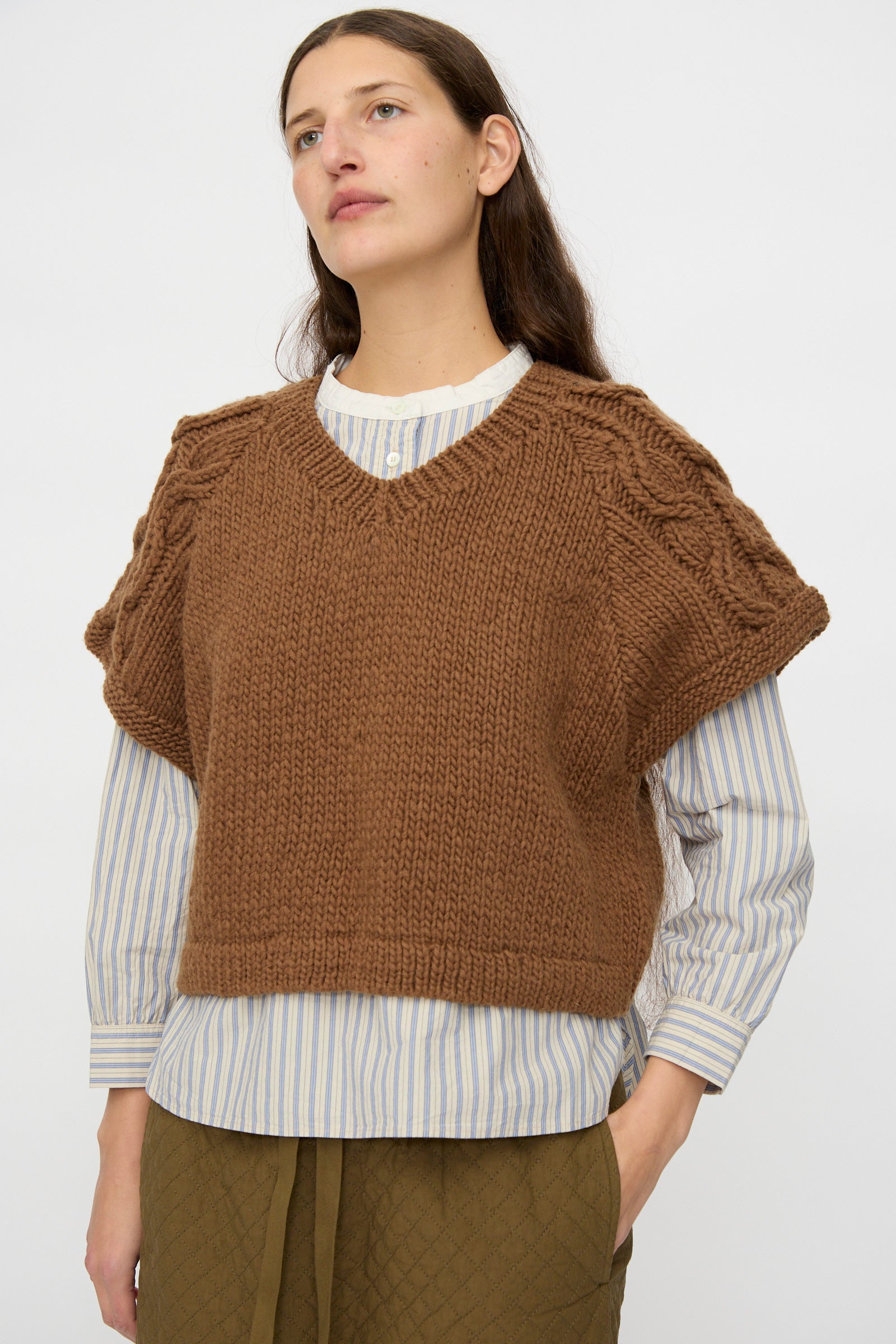 A woman wears the nest Robe Peruvian Wool Raglan Cable Knit Vest in Camel over a blue and white striped shirt and green quilted pants, standing against a plain light background.