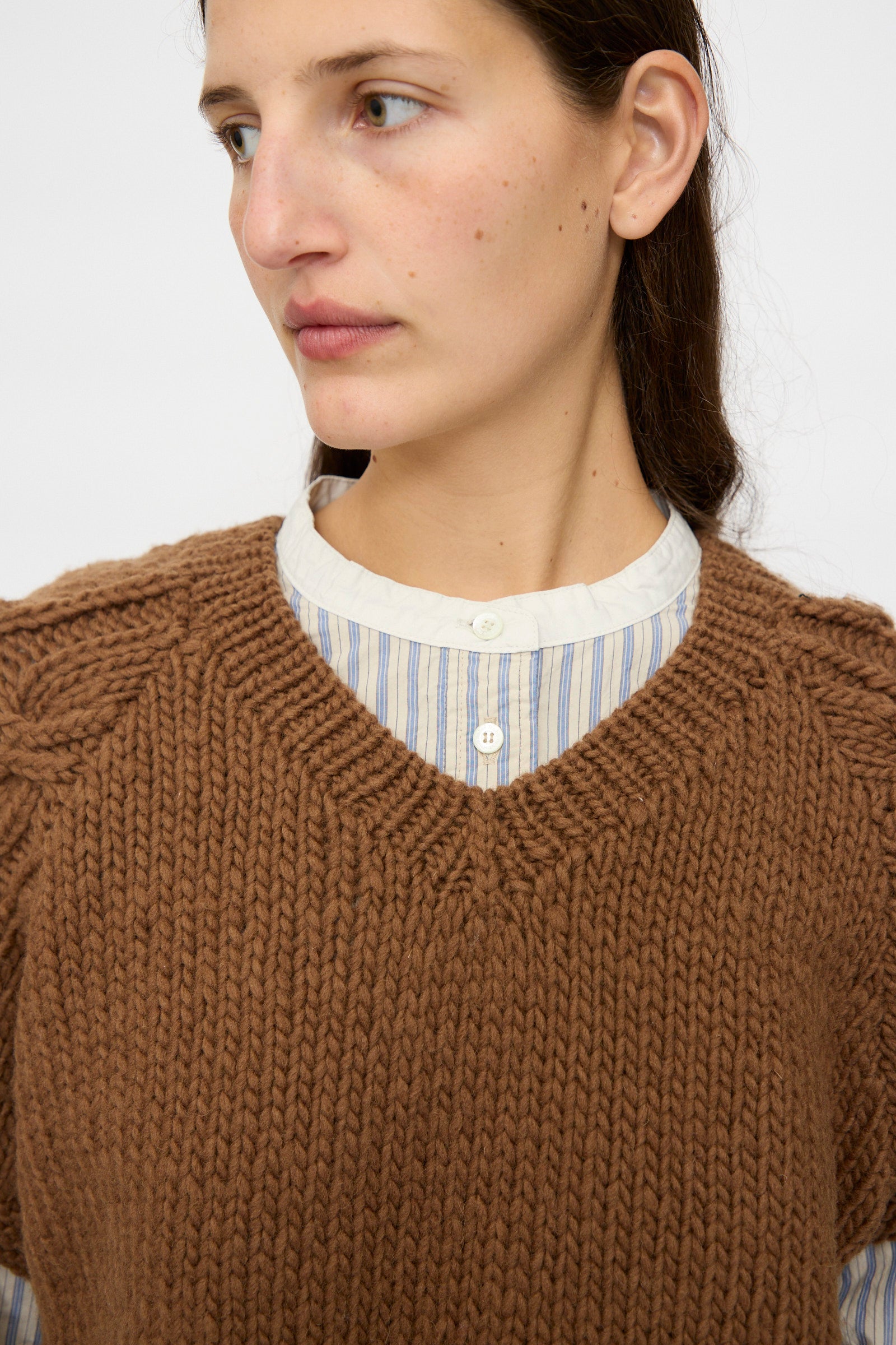 A person wears the nest Robe Peruvian Wool Raglan Cable Knit Vest in Camel over a blue and white striped button-up shirt, looking slightly to the side.