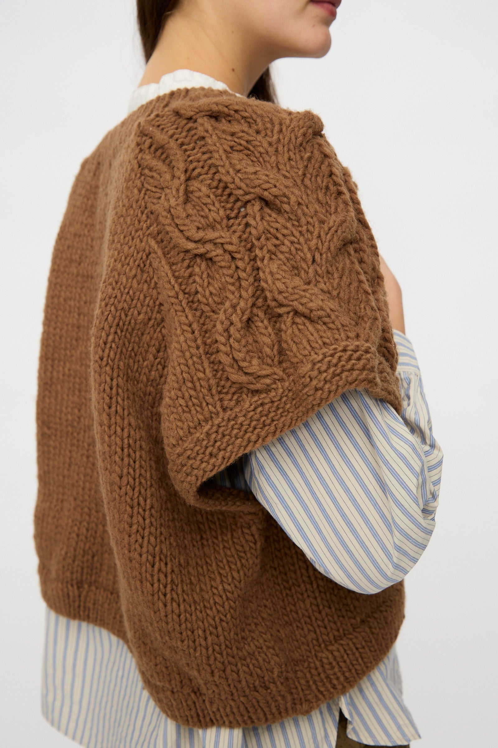A person is seen from the side wearing nest Robe's Peruvian Wool Raglan Cable Knit Vest in Camel over a blue and white striped shirt.