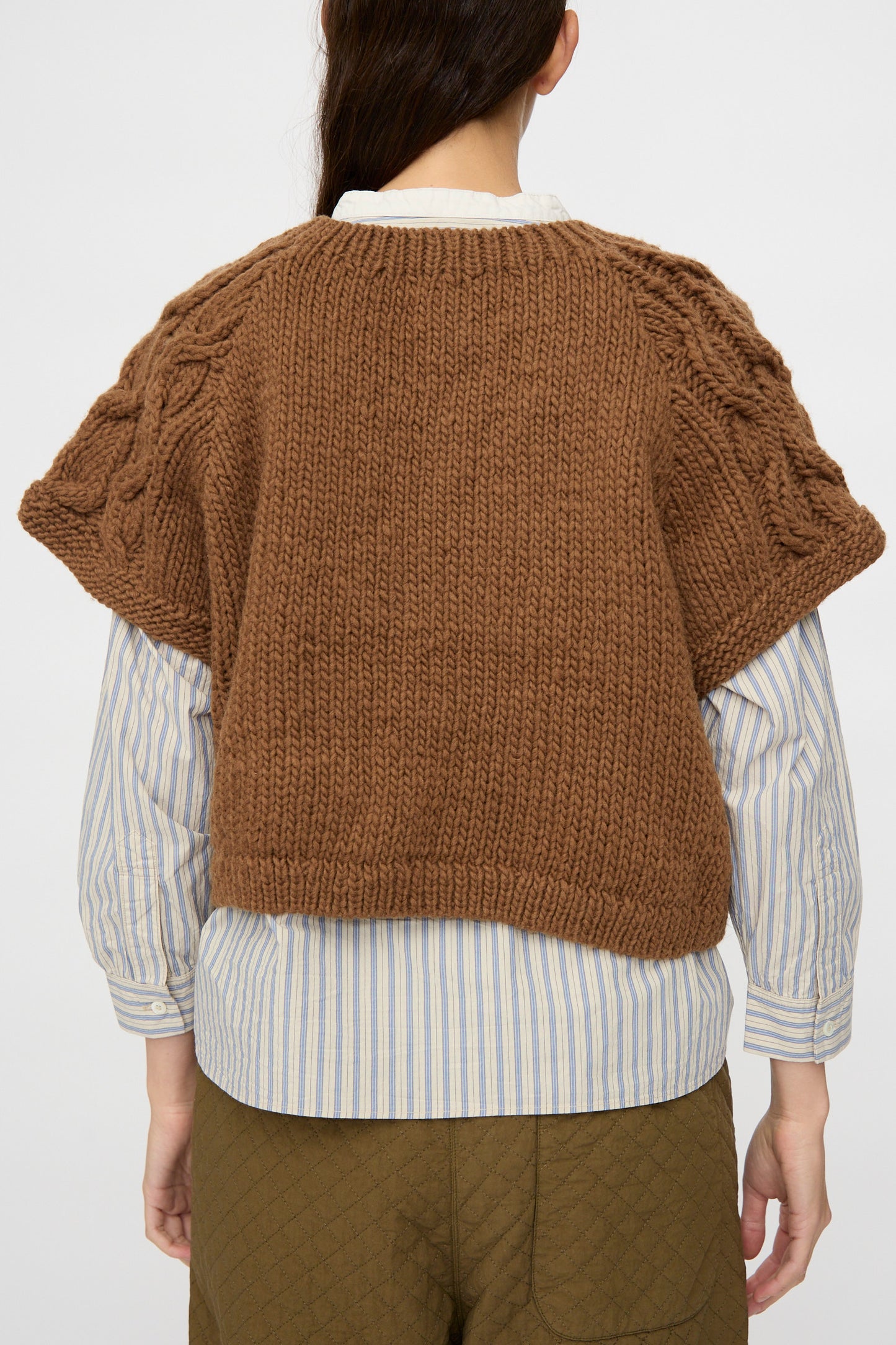 A person shown from the back is wearing the nest Robe Peruvian Wool Raglan Cable Knit Vest in Camel over a blue-and-white striped shirt and olive quilted pants.