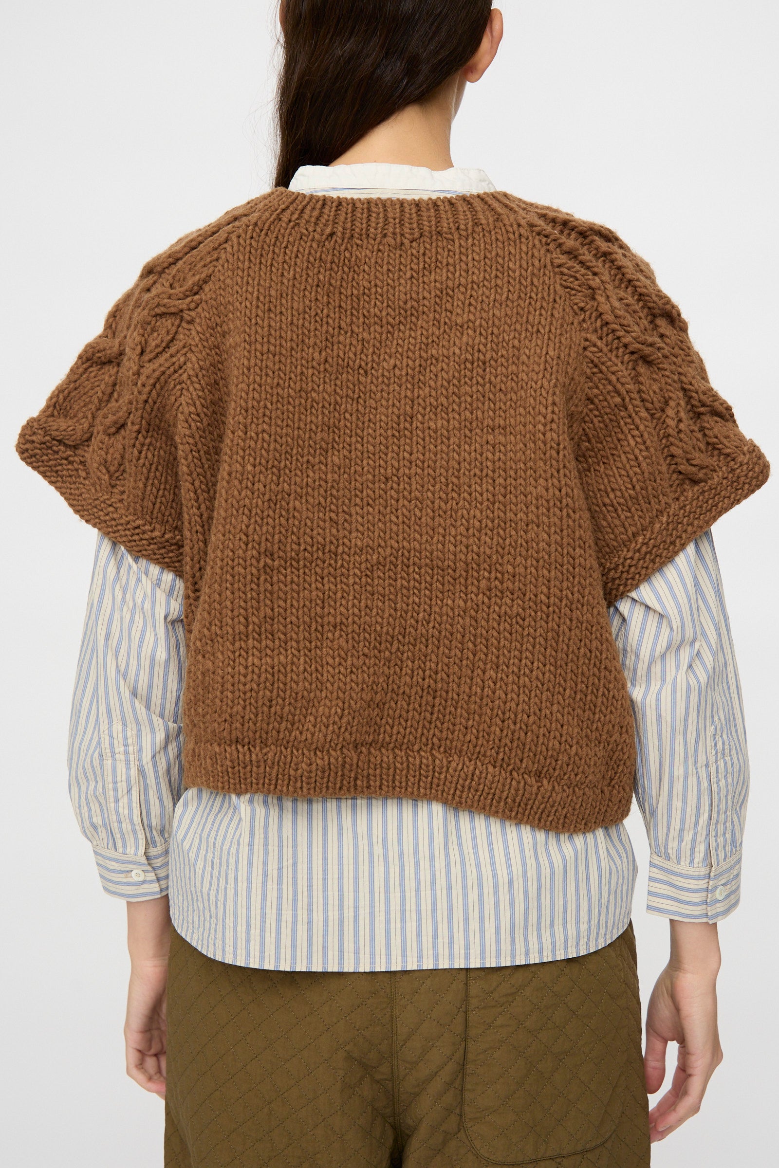 A person shown from the back is wearing the nest Robe Peruvian Wool Raglan Cable Knit Vest in Camel over a blue-and-white striped shirt and olive quilted pants.