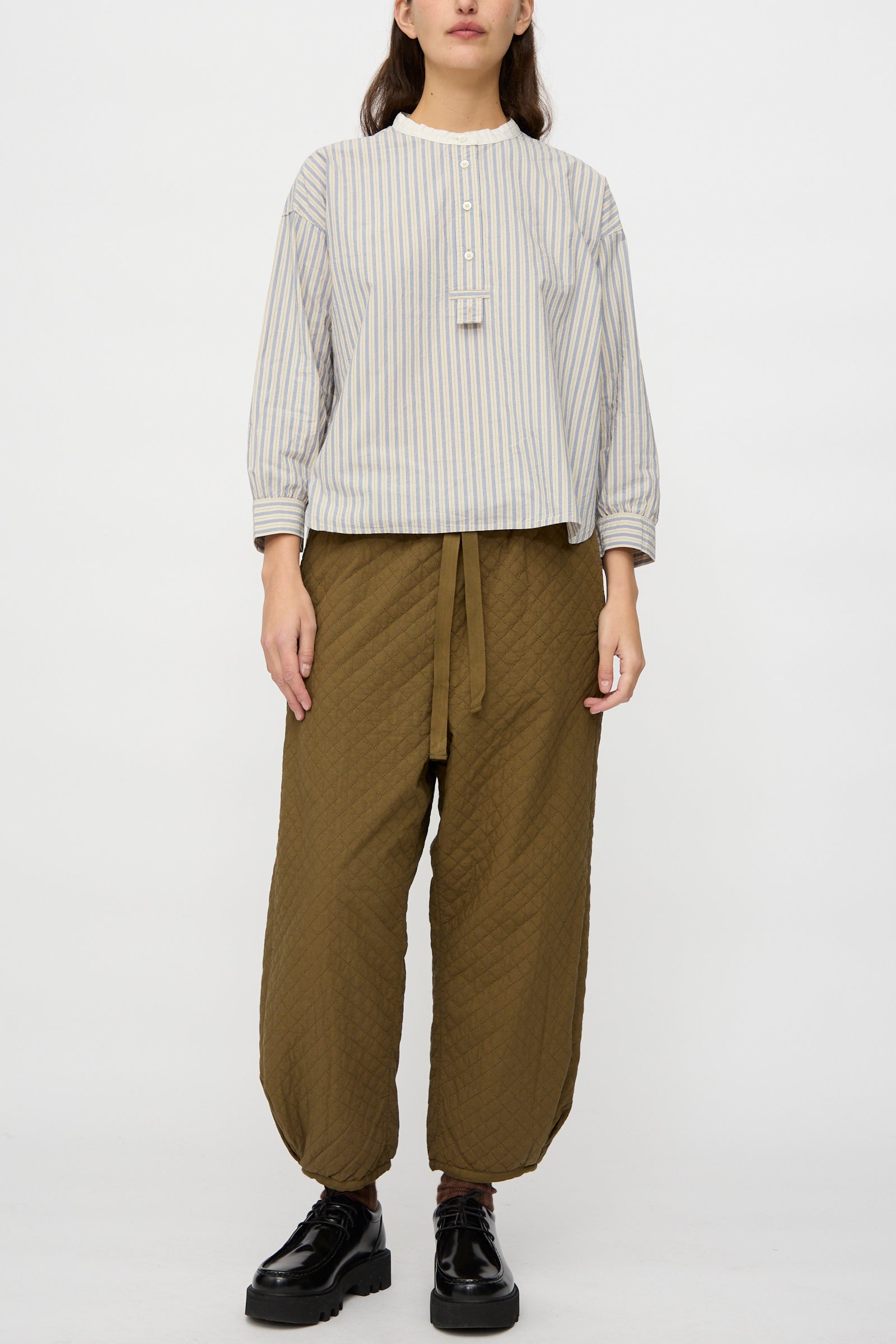 A person wears nest Robe's Quilted Drawstring Trouser in Olive with a light-colored, long-sleeve striped button-up shirt and black shoes, standing against a plain white background.