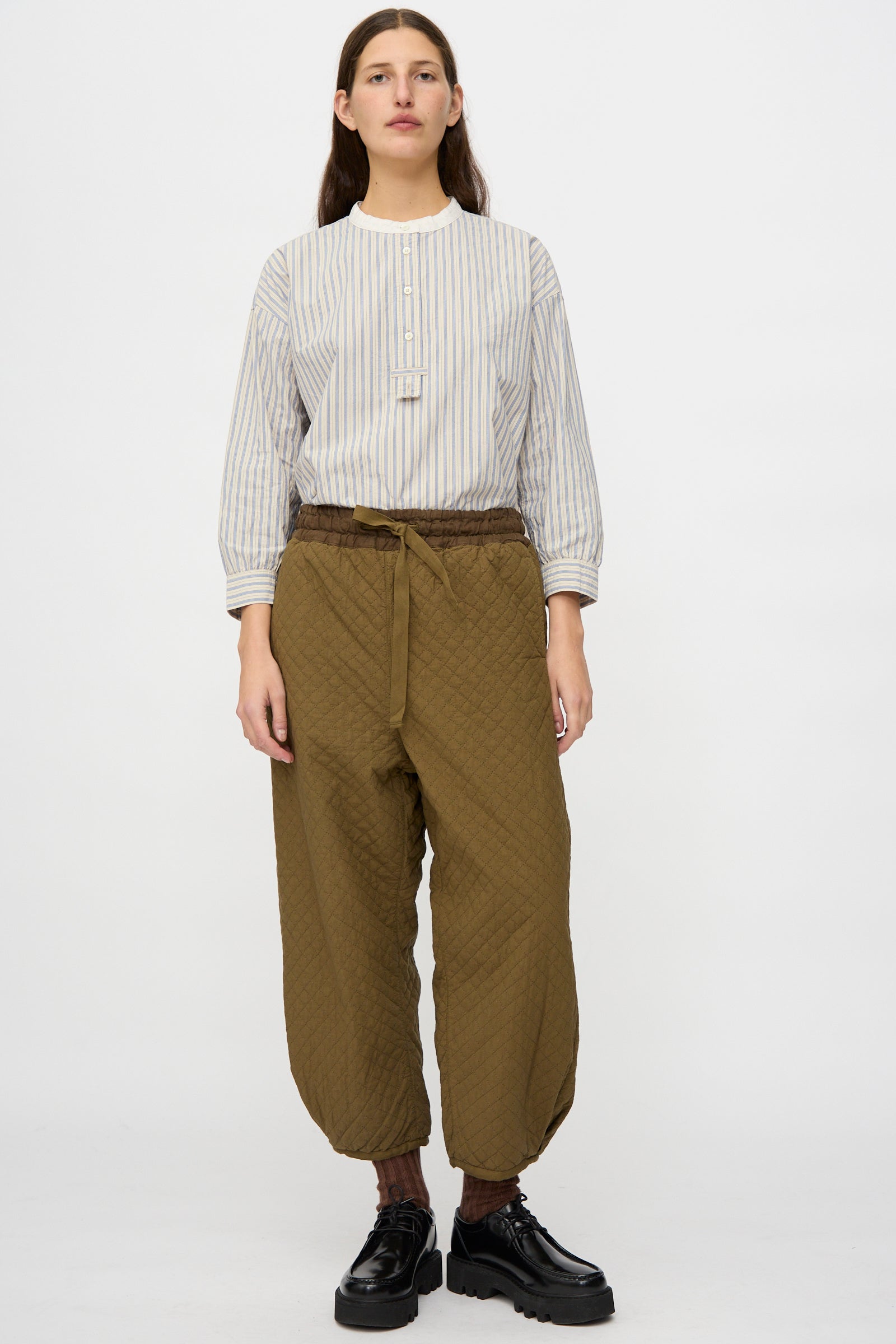 A person stands facing forward, wearing a striped long-sleeve shirt, nest Robe Quilted Drawstring Trouser in Olive, and black platform shoes against a plain white background.