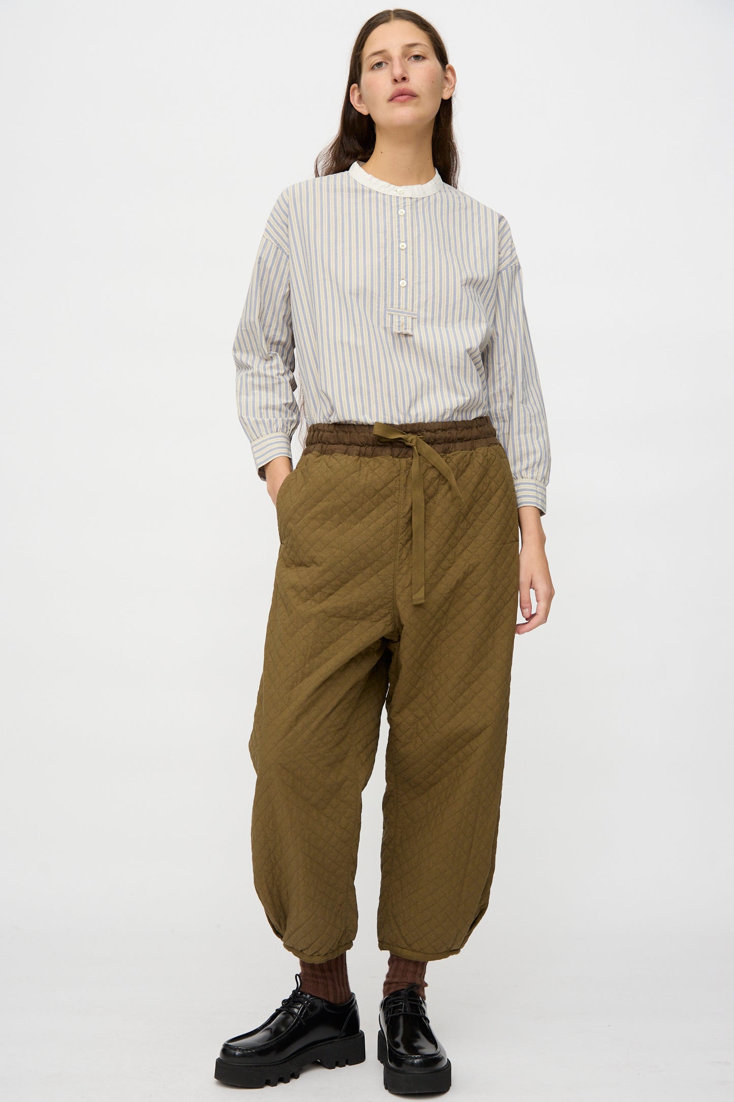 A person stands against a plain white background wearing a striped long-sleeve shirt, nest Robe's Quilted Drawstring Trouser in Olive, brown socks, and black platform shoes.