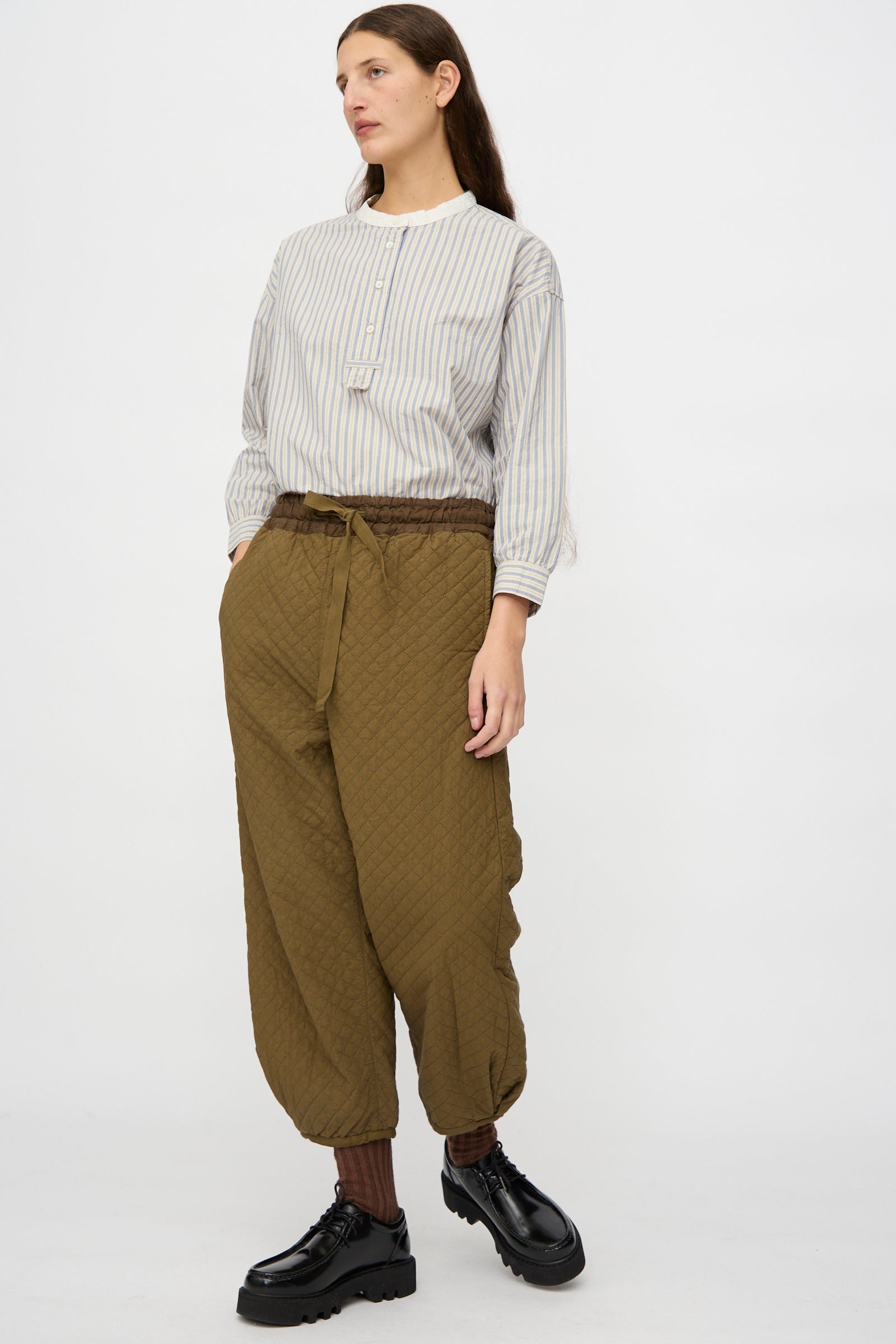 A person stands against a plain background wearing a striped long-sleeve shirt, brown socks, black platform shoes, and the nest Robe Quilted Drawstring Trouser in Olive.