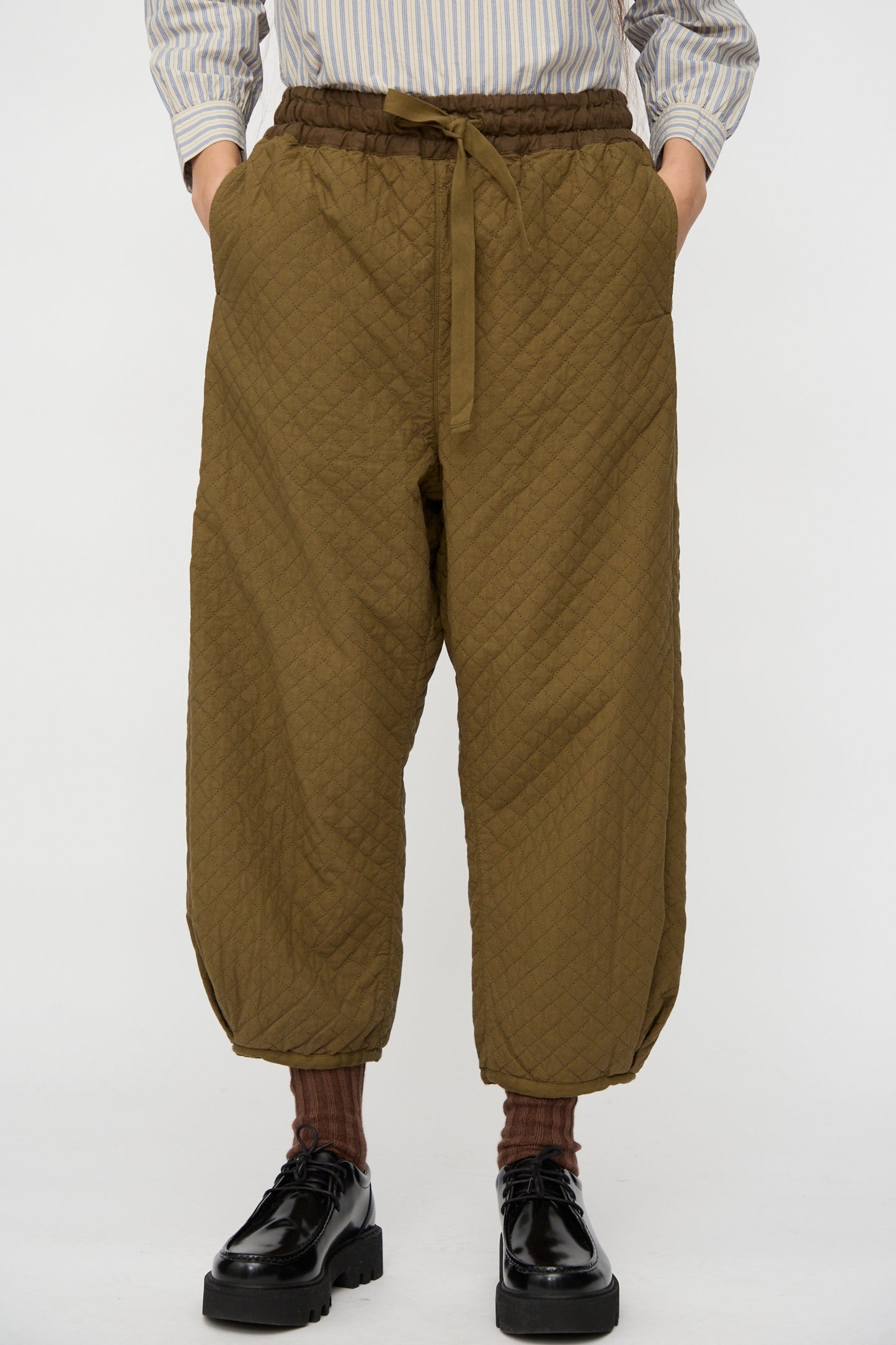 A person wears nest Robe’s Quilted Drawstring Trouser in Olive with a striped shirt, brown socks, and black shoes. Hands are casually tucked into the trouser pockets.