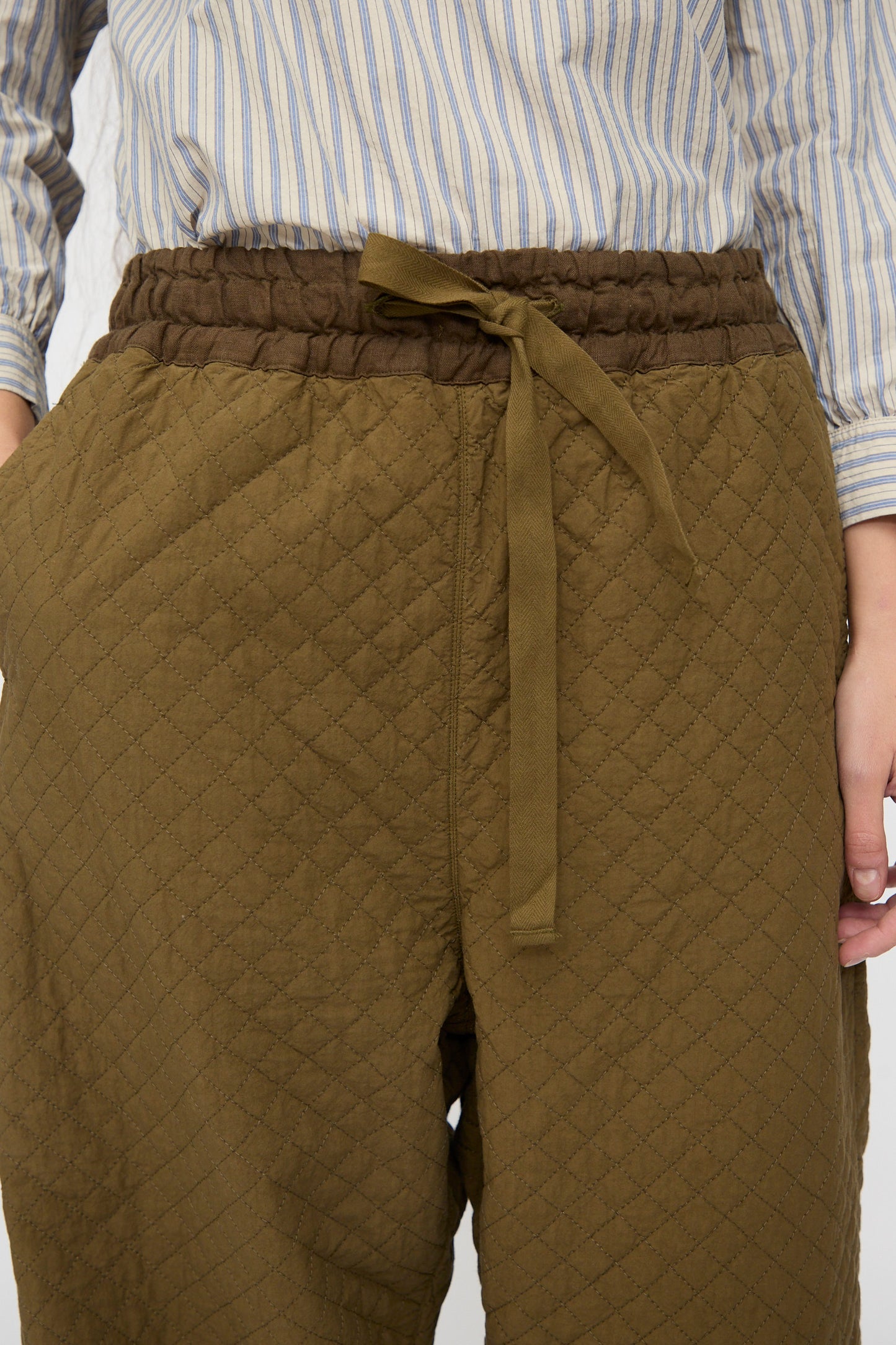 Someone wears the nest Robe Quilted Drawstring Trouser in Olive with an elastic waistband, paired with a blue and white striped shirt.