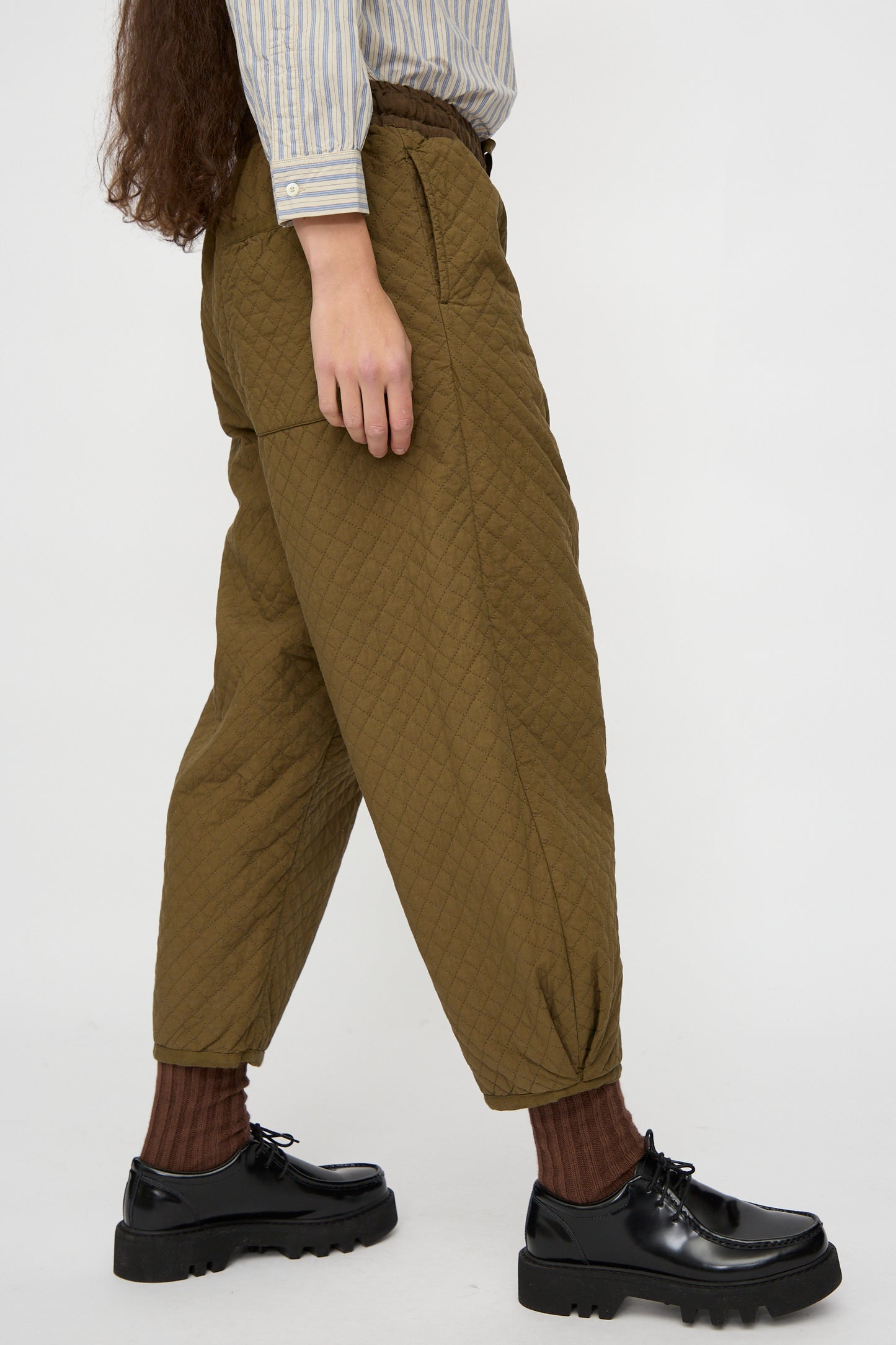 A person wears the nest Robe Quilted Drawstring Trouser in Olive, paired with a striped shirt, brown socks, and black platform shoes against a plain white backdrop.