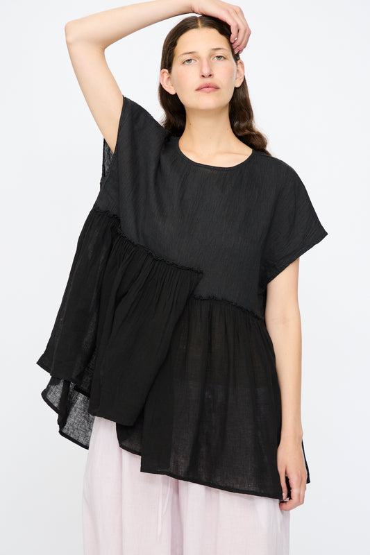A person wears the nest Robe Ramie Crepe Linen Gauze Blouse in Black, featuring ruffled layers and wide short sleeves, paired with light-colored pants and standing against a plain background.