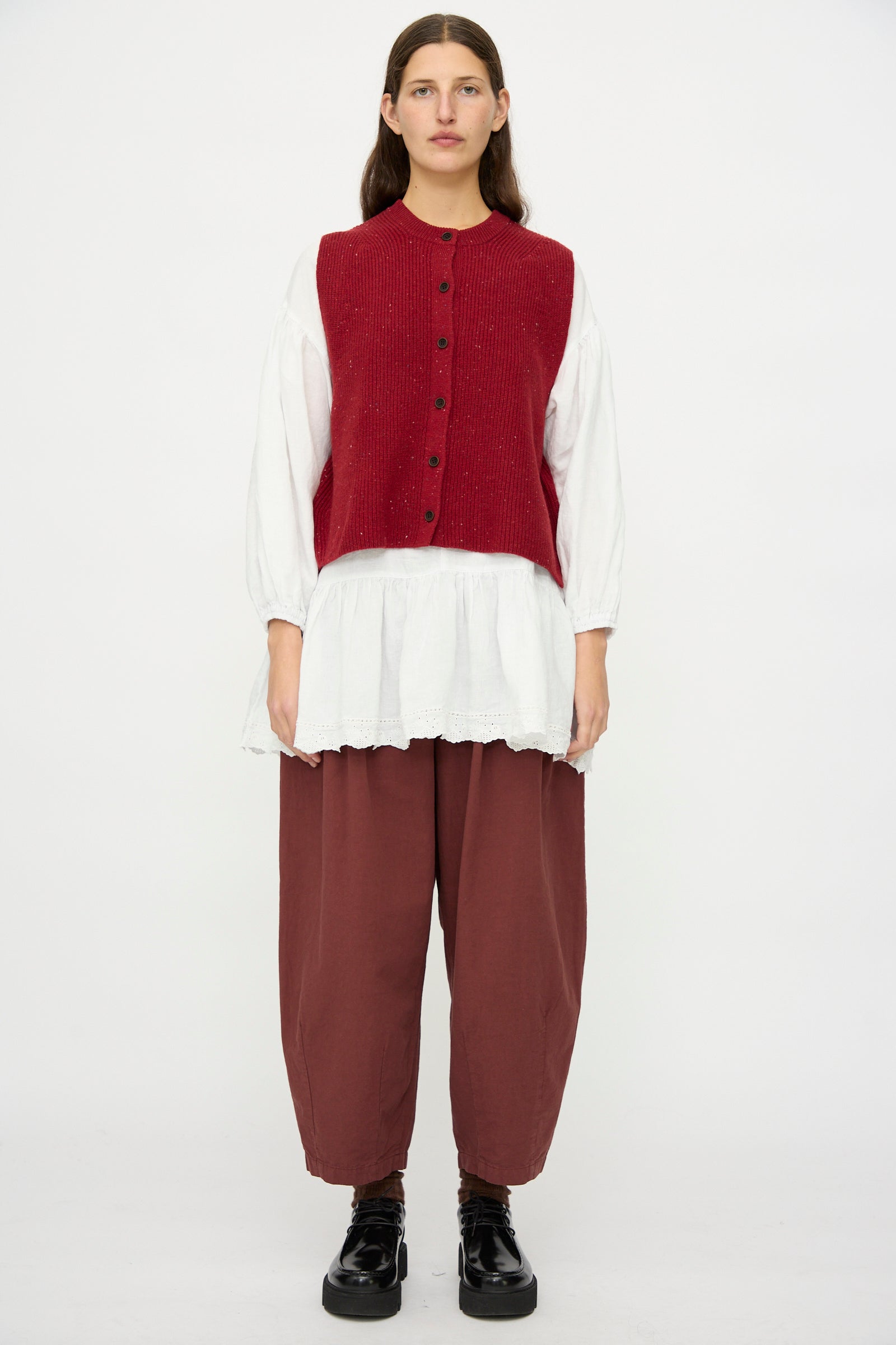 A person stands facing forward, wearing the nest Robe Rib Knit Buttoned Vest in Wine over a loose white blouse, brown wide-leg pants, and black shoes against a plain white background.