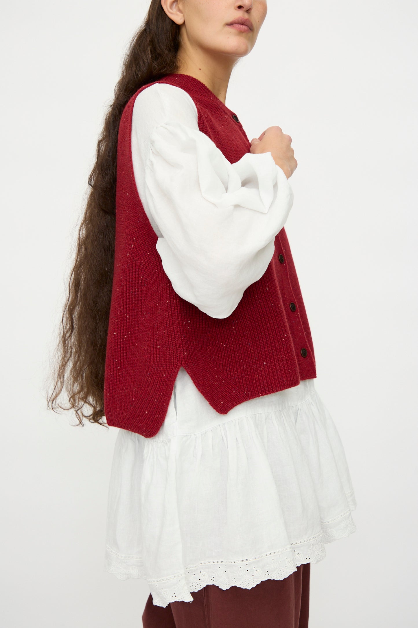 A model in the nest Robe Rib Knit Buttoned Vest in Wine, layered over a white puff-sleeve blouse with a ruffled hem, is shown from the side against a plain background.