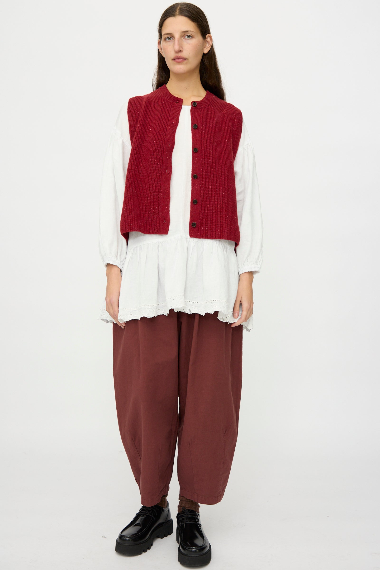 A woman stands before a white background wearing the nest Robe Rib Knit Buttoned Vest in Wine, a white long-sleeve blouse, maroon loose pants, and black shoes.