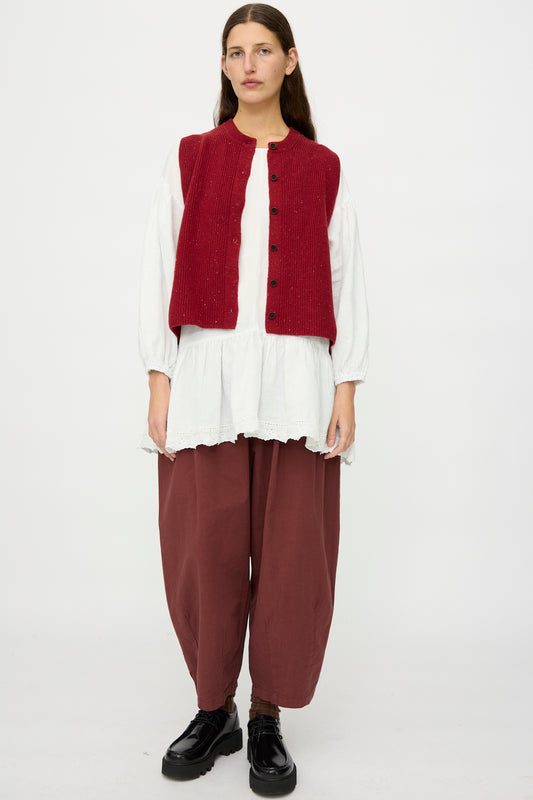A woman stands before a white background wearing the nest Robe Rib Knit Buttoned Vest in Wine, a white long-sleeve blouse, maroon loose pants, and black shoes.