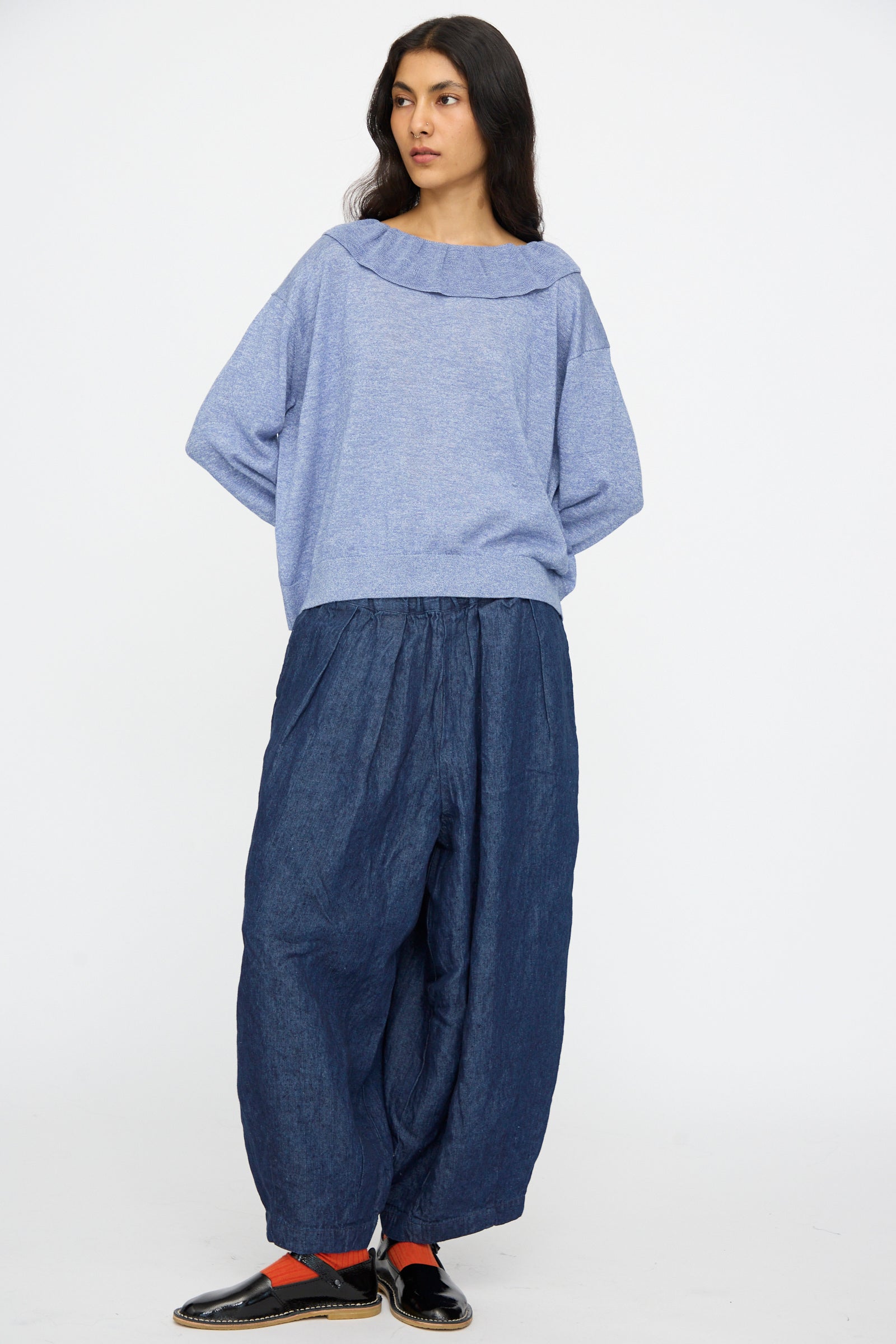 A person stands against a white background, wearing the nest Robe Ruffle Collar Knit Pullover in Blue, paired with baggy dark blue pants, black slip-on shoes, and orange socks.