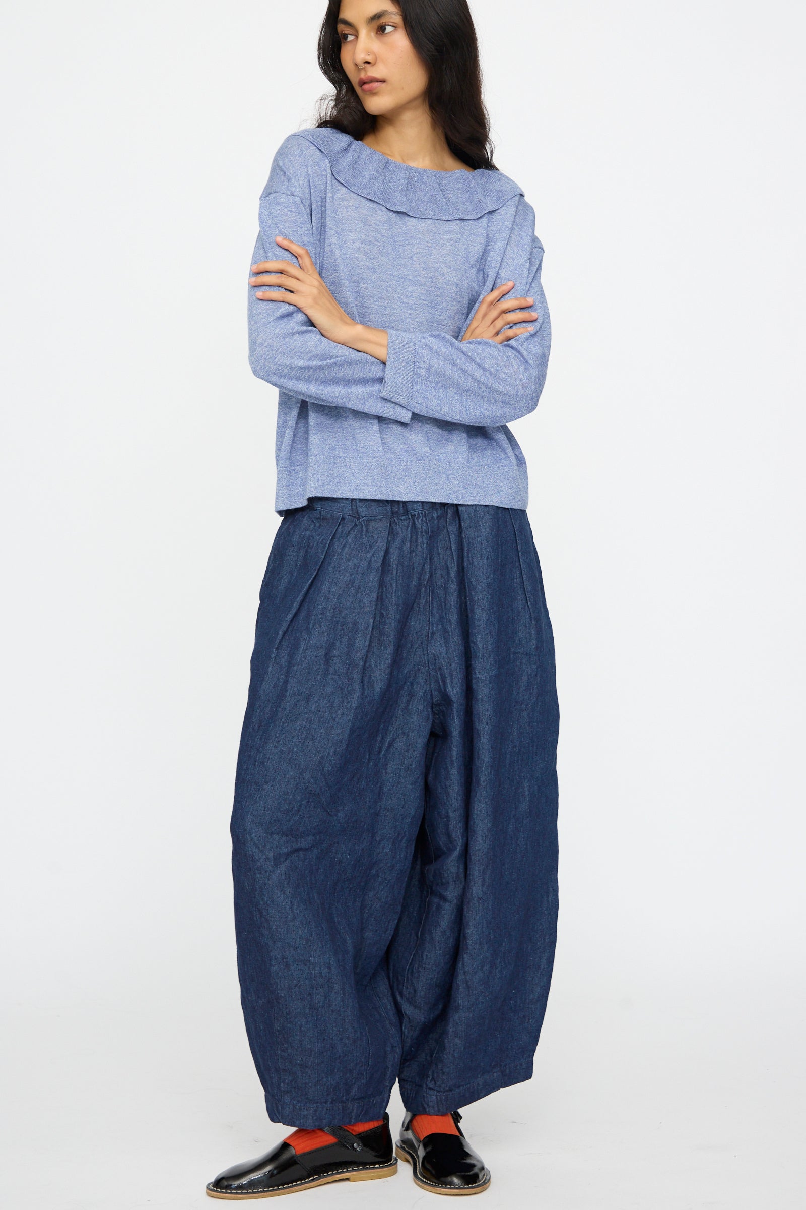 A person stands with arms crossed, wearing the nest Robe Ruffle Collar Knit Pullover in Blue, loose dark blue pants, black shoes, and red socks against a plain white background.