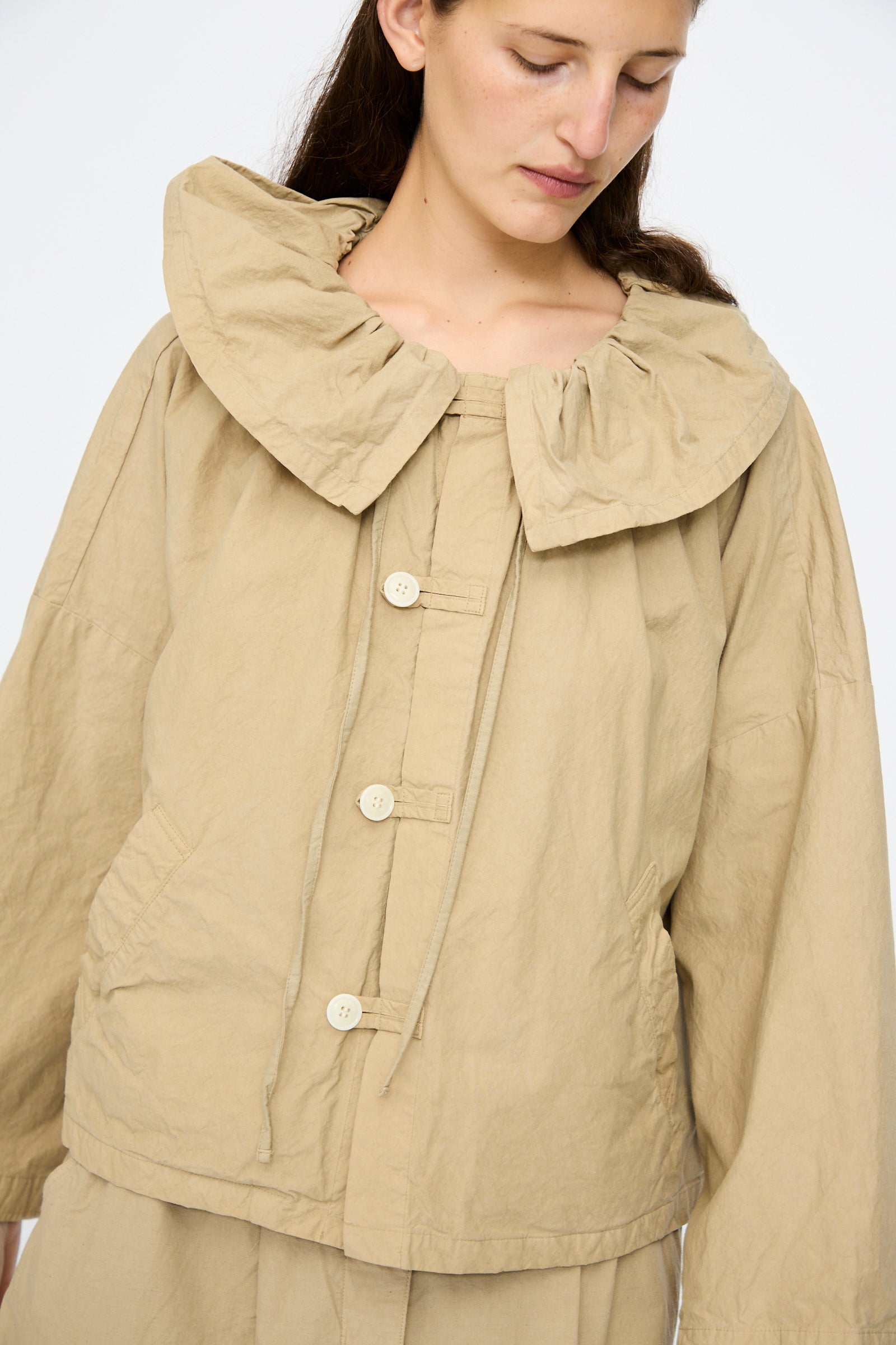 A woman wears the nest Robe Super Water-Repellent Supima Cotton Leaf Collar Jacket in beige, featuring a boxy fit and ruffled collar, paired with matching beige pants.