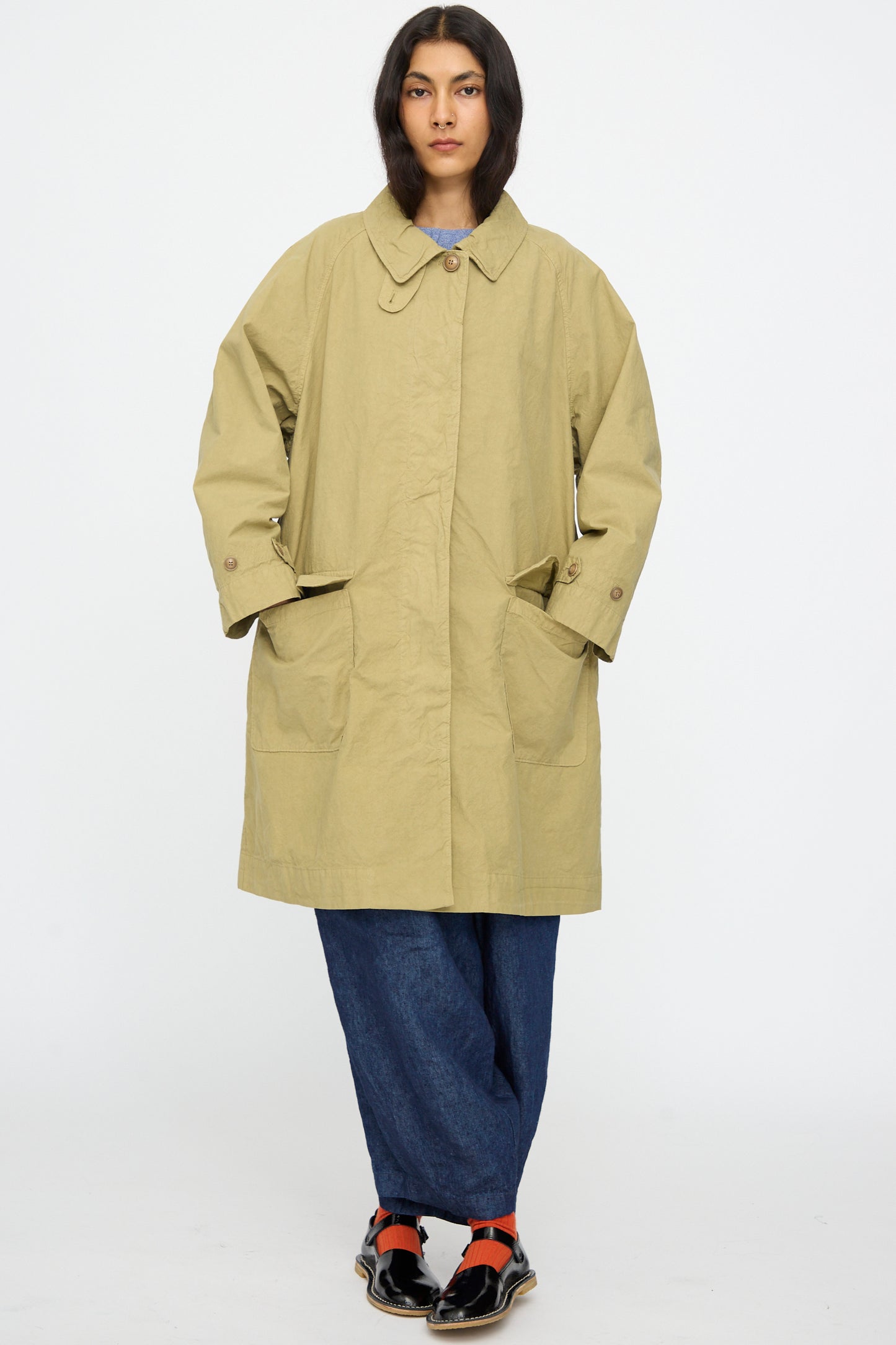 A person stands against a white background wearing the nest Robe Supima Cotton Topcoat in Green, blue pants, black shoes, and red socks.