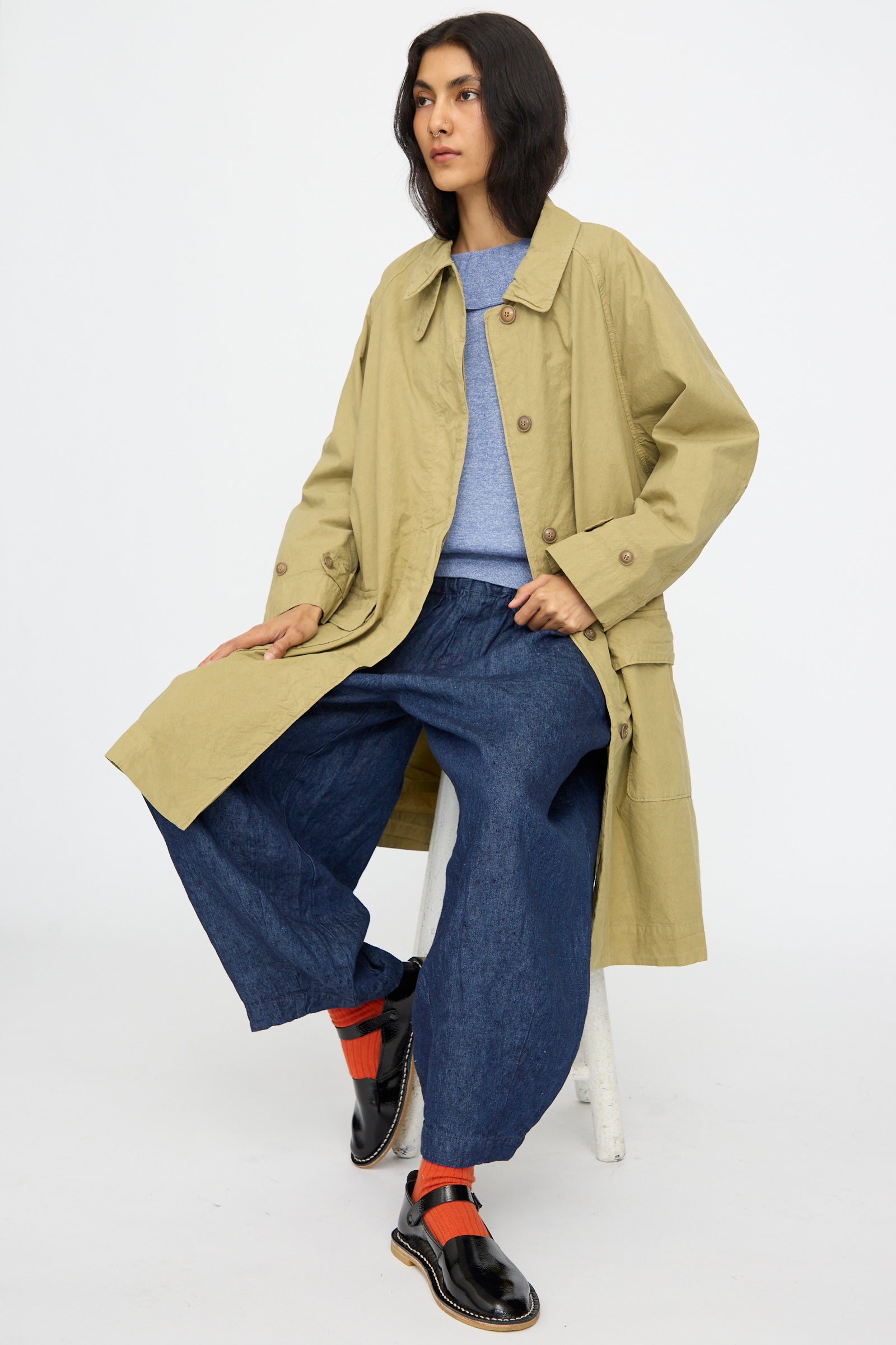 A person sits on a stool against a plain white background, wearing nest Robe's Supima Cotton Topcoat in Green, paired with a blue sweater, loose blue pants, black shoes, and red socks.
