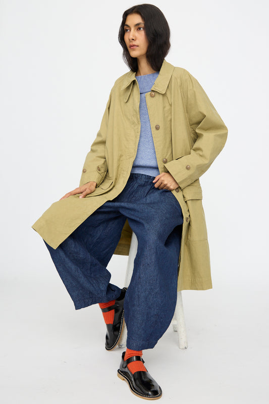 A person sits on a stool against a plain white background, wearing nest Robe's Supima Cotton Topcoat in Green, paired with a blue sweater, loose blue pants, black shoes, and red socks.