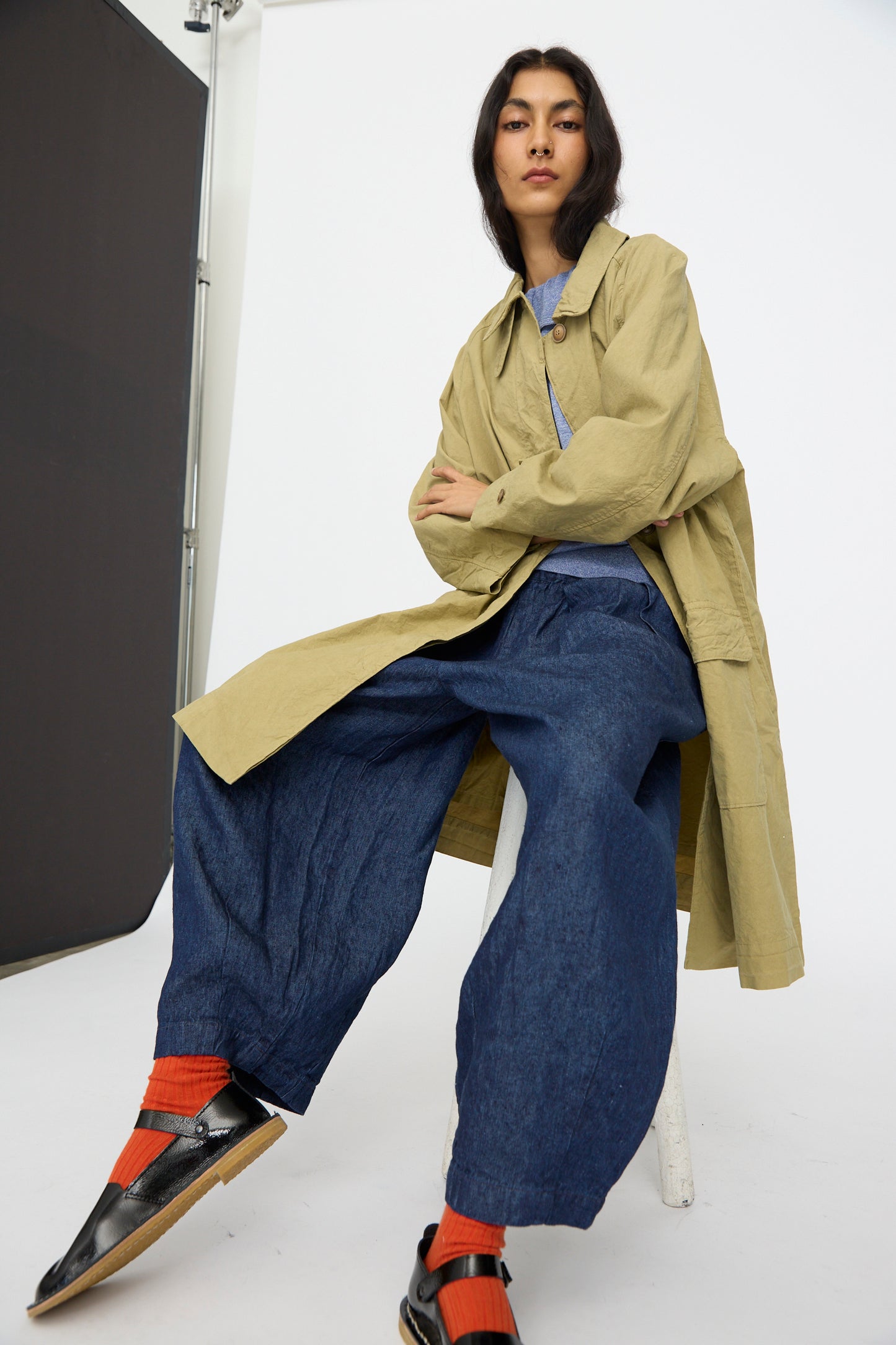 A person sits on a stool against a plain studio backdrop, wearing the nest Robe Supima Cotton Topcoat in Green (made in Japan), blue wide-leg pants, orange socks, and black shoes.