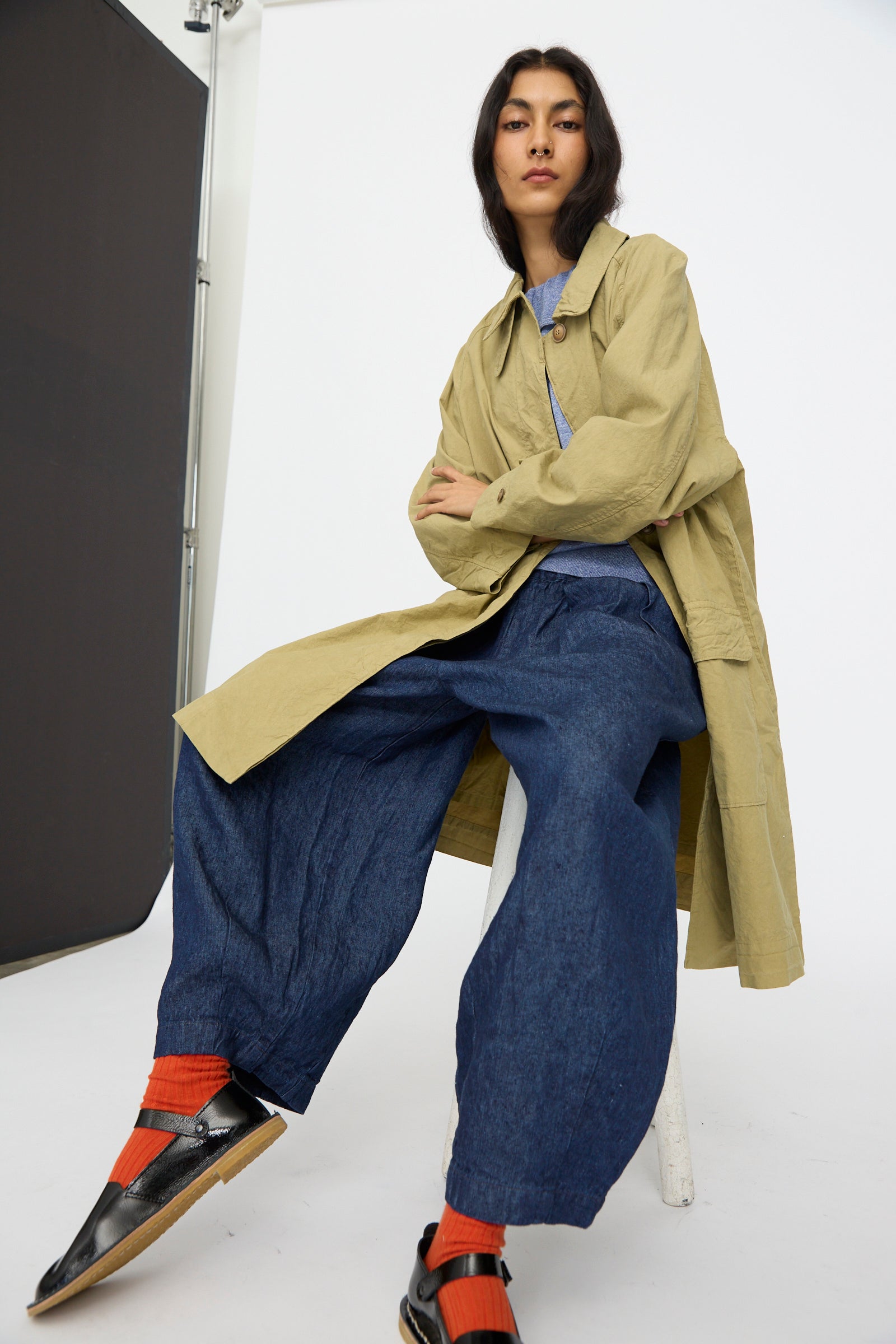 A person sits on a stool against a plain studio backdrop, wearing the nest Robe Supima Cotton Topcoat in Green (made in Japan), blue wide-leg pants, orange socks, and black shoes.