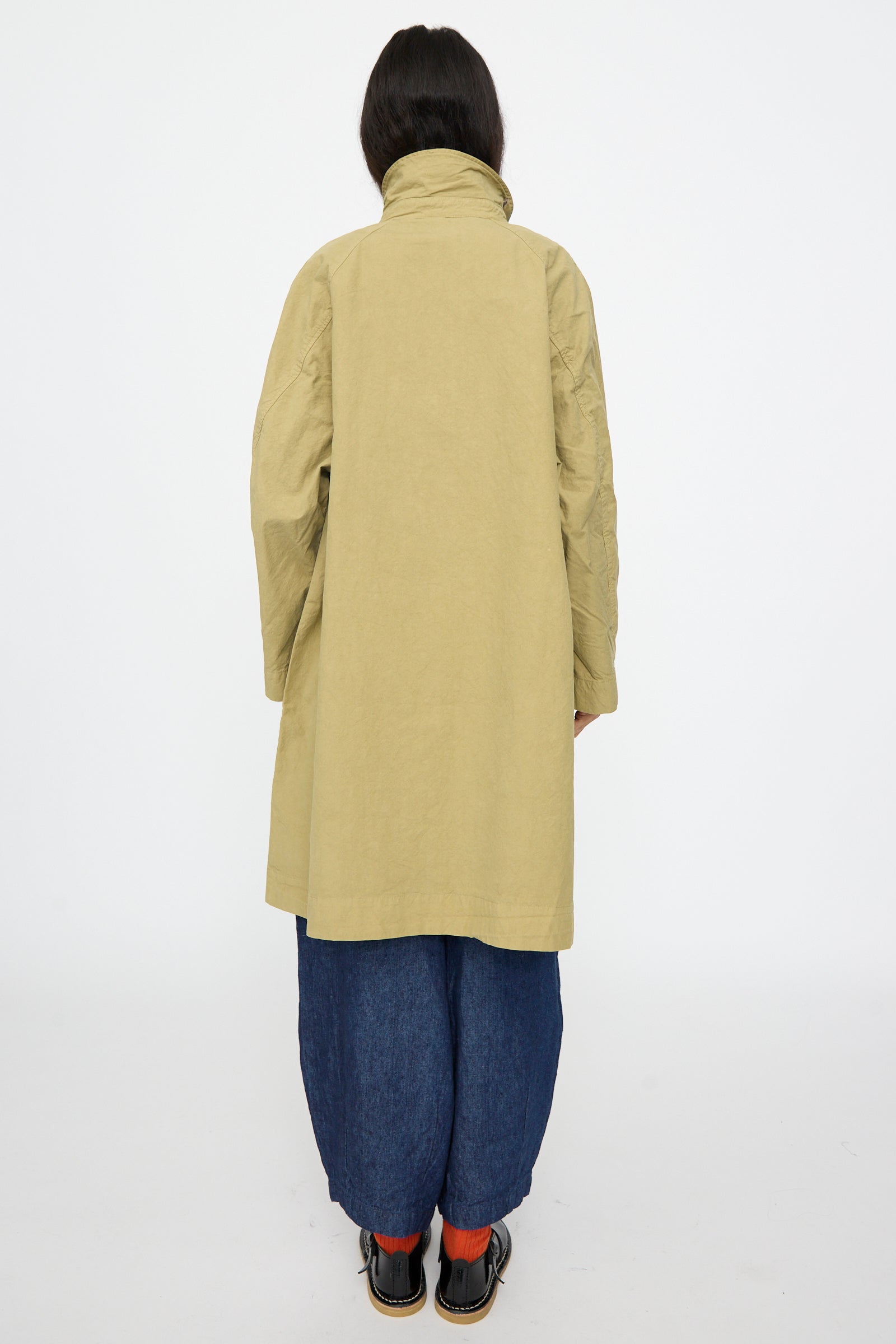 A person faces away from the camera wearing nest Robe's Supima Cotton Topcoat in Green, paired with wide dark blue pants, orange socks, and black shoes, all set against a plain white background. The coat is made in Japan for added quality.