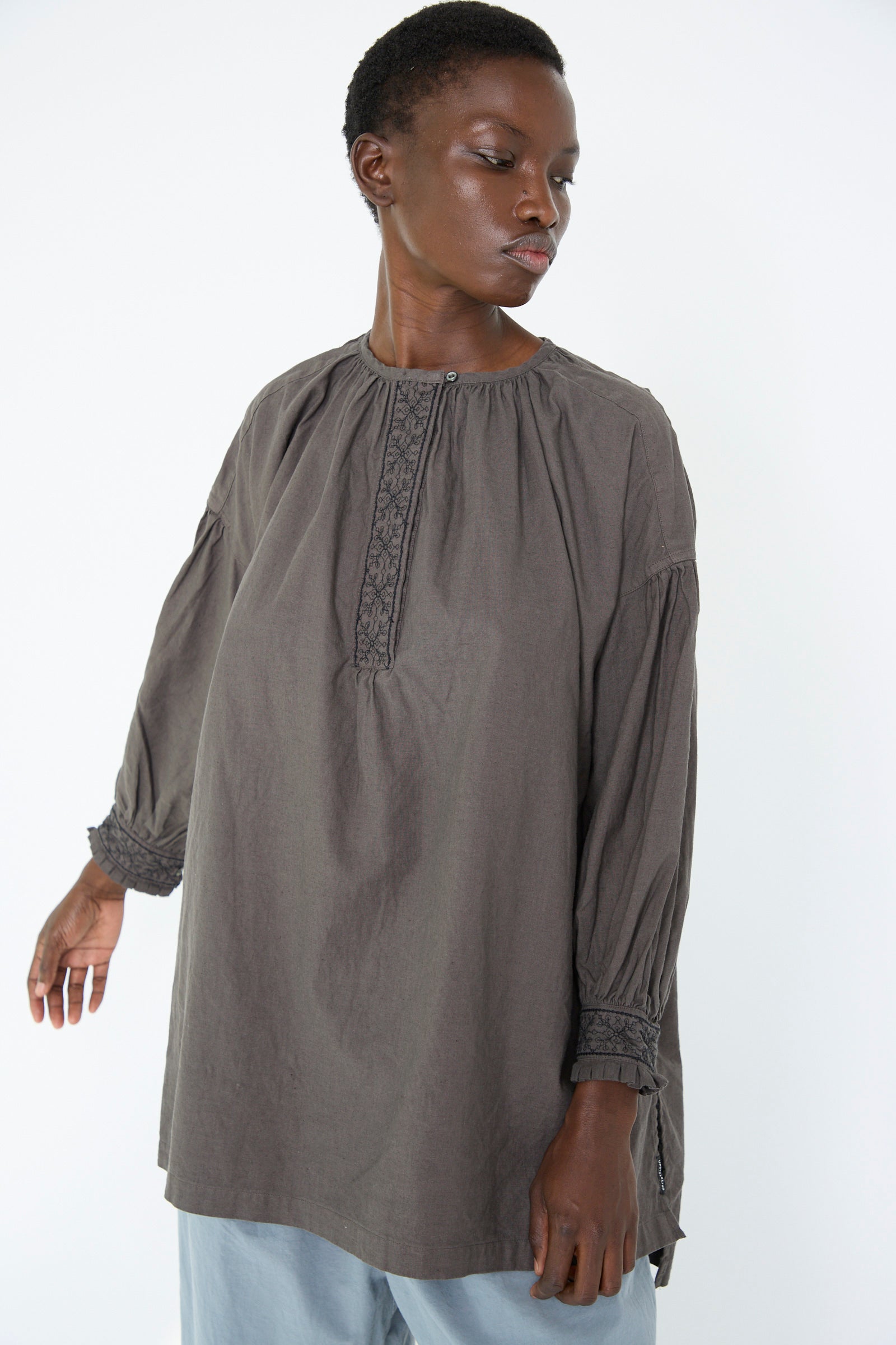 A woman dressed in an UpcycleLino Cotton Linen Embroidered Tunic in Grey by nest Robe, is standing against a plain white background and looking away.