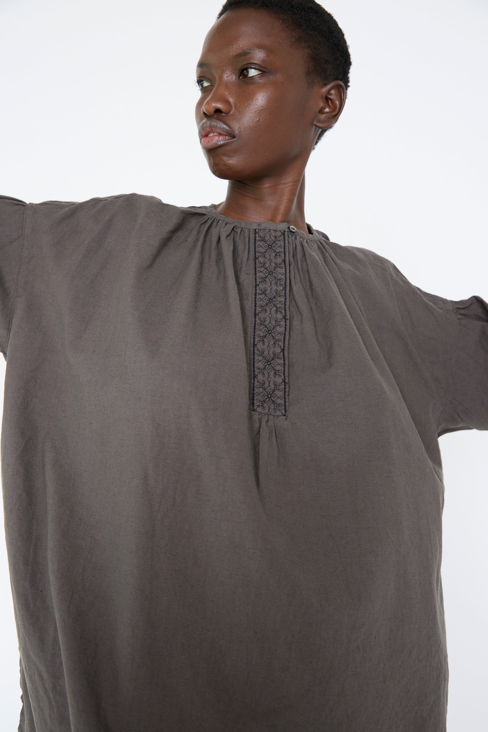 A woman wearing the UpcycleLino Cotton Linen Embroidered Tunic in Grey by nest Robe, featuring blackwork embroidery stands with arms slightly raised against a plain background.