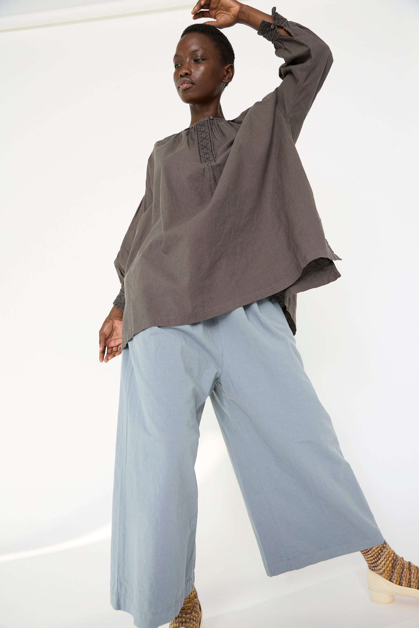 A woman wearing the UpcycleLino Cotton Linen Embroidered Tunic in Grey by nest Robe, paired with light blue wide-leg pants, poses against a plain background.