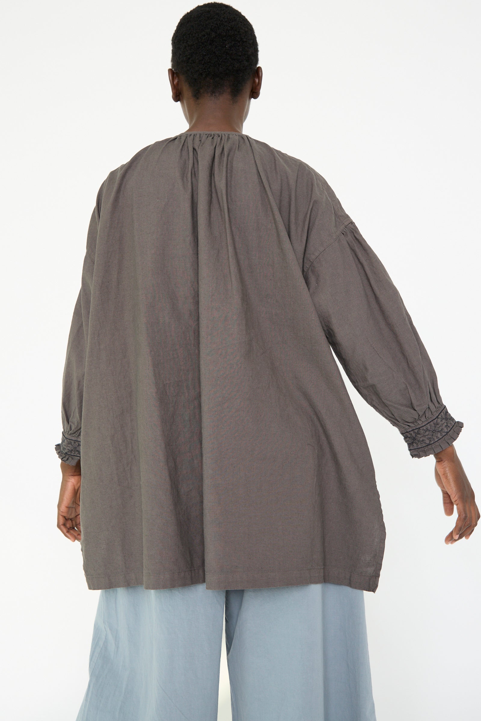 A woman wearing the UpcycleLino Cotton Linen Embroidered Tunic in Grey by nest Robe and light blue pants, facing away from the camera.