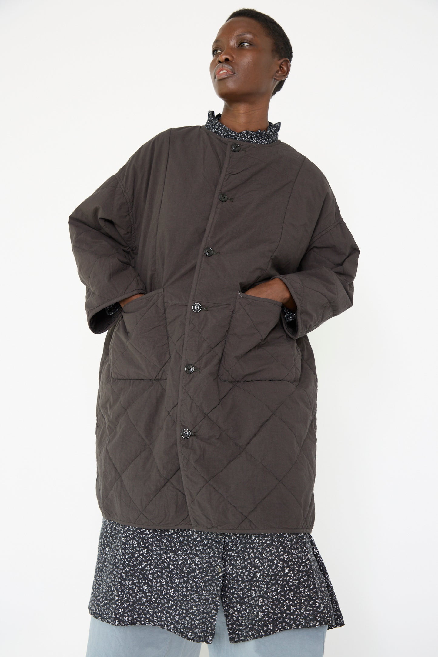 A person is wearing the UpcycleLino Reversible Quilted Coat in Ink Black by nest Robe, with hands tucked into the pockets, over a floral patterned top and light blue pants, standing against a plain background.