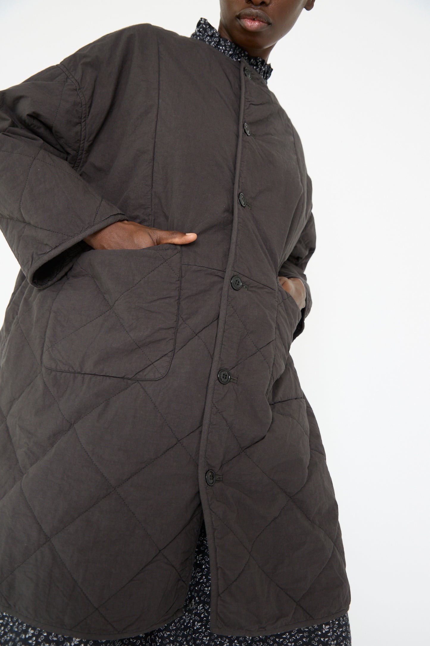 A woman wearing the UpcycleLino Reversible Quilted Coat in Ink Black by nest Robe over a patterned dress, with her hands in the pockets.