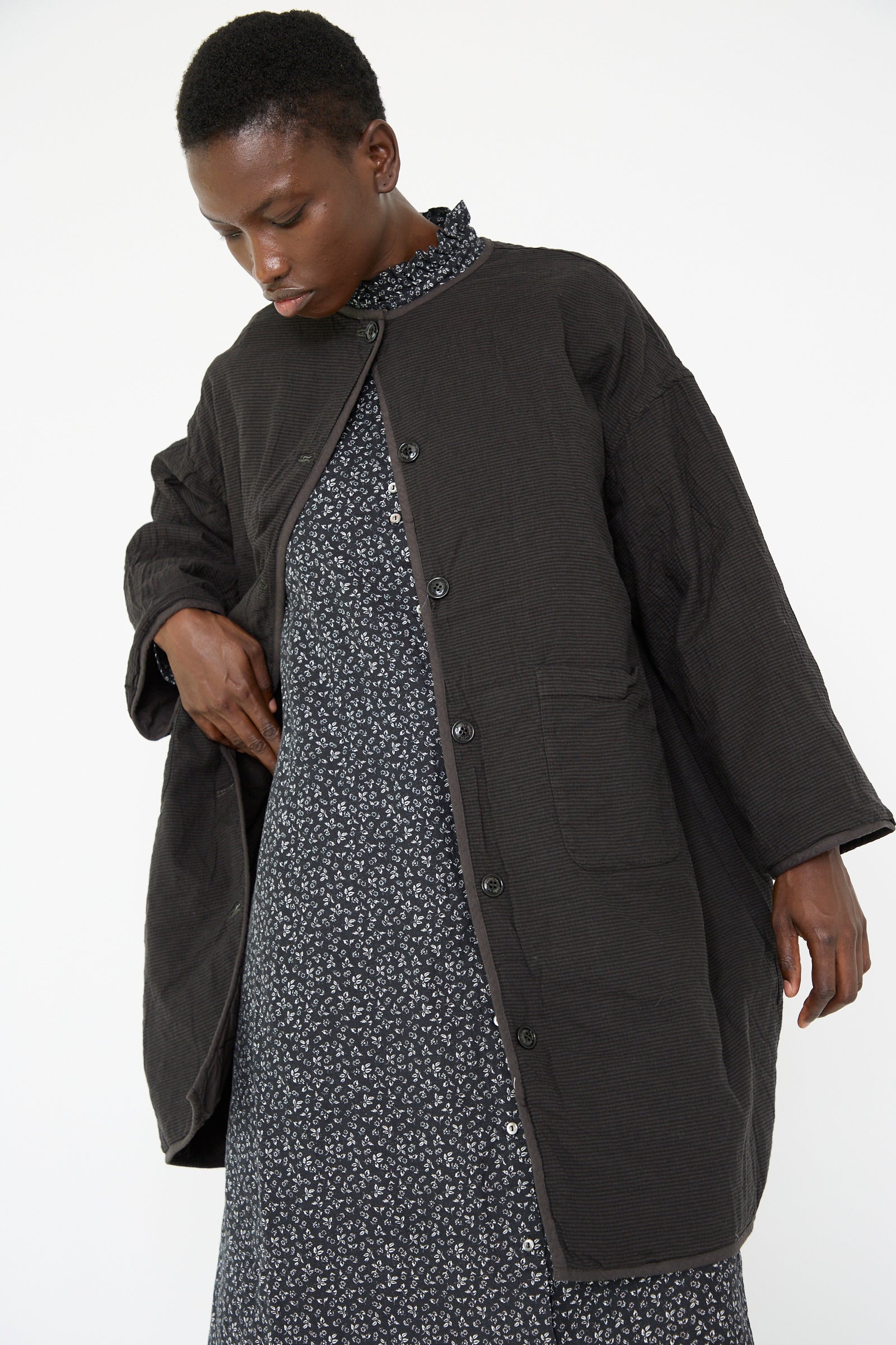 A woman wearing a nest Robe UpcycleLino Reversible Quilted Coat in Ink Black over a patterned dress, gazing downward.