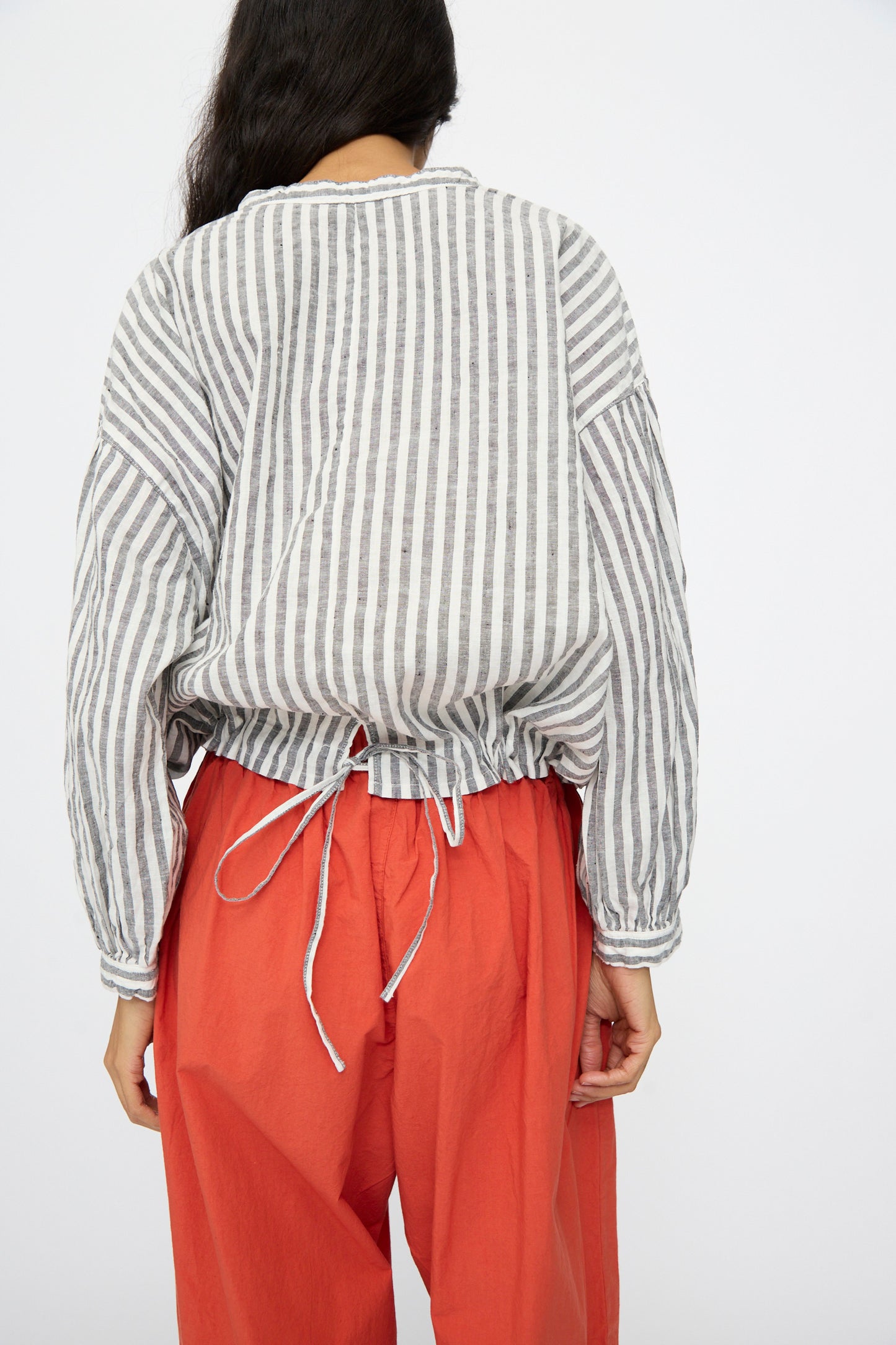 A person faces away, wearing the nest Robe Upcyclelino Ribbon Two-Way Blouse in Stripe, featuring a gray and white pattern with an adjustable cinched hem, paired with loose orange pants.