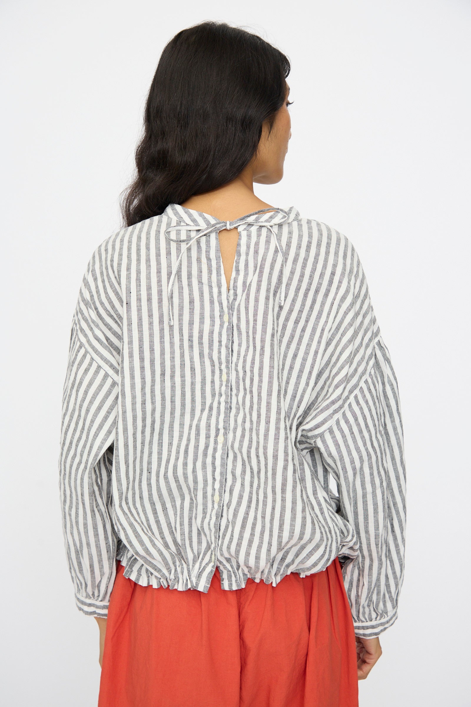 A person with long dark hair faces away, wearing the nest Robe Upcyclelino Ribbon Two-Way Blouse in Stripe, a loose linen-cotton top with back tie, button details, adjustable cinched hem, and paired with a red skirt.