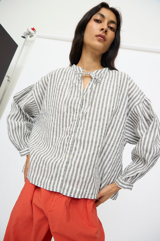 A person wears the nest Robe Upcyclelino Ribbon Two-Way Blouse in Stripe—gray and white, linen cotton blend—styled with bright red pants and poses with one hand on hip against a plain light background.