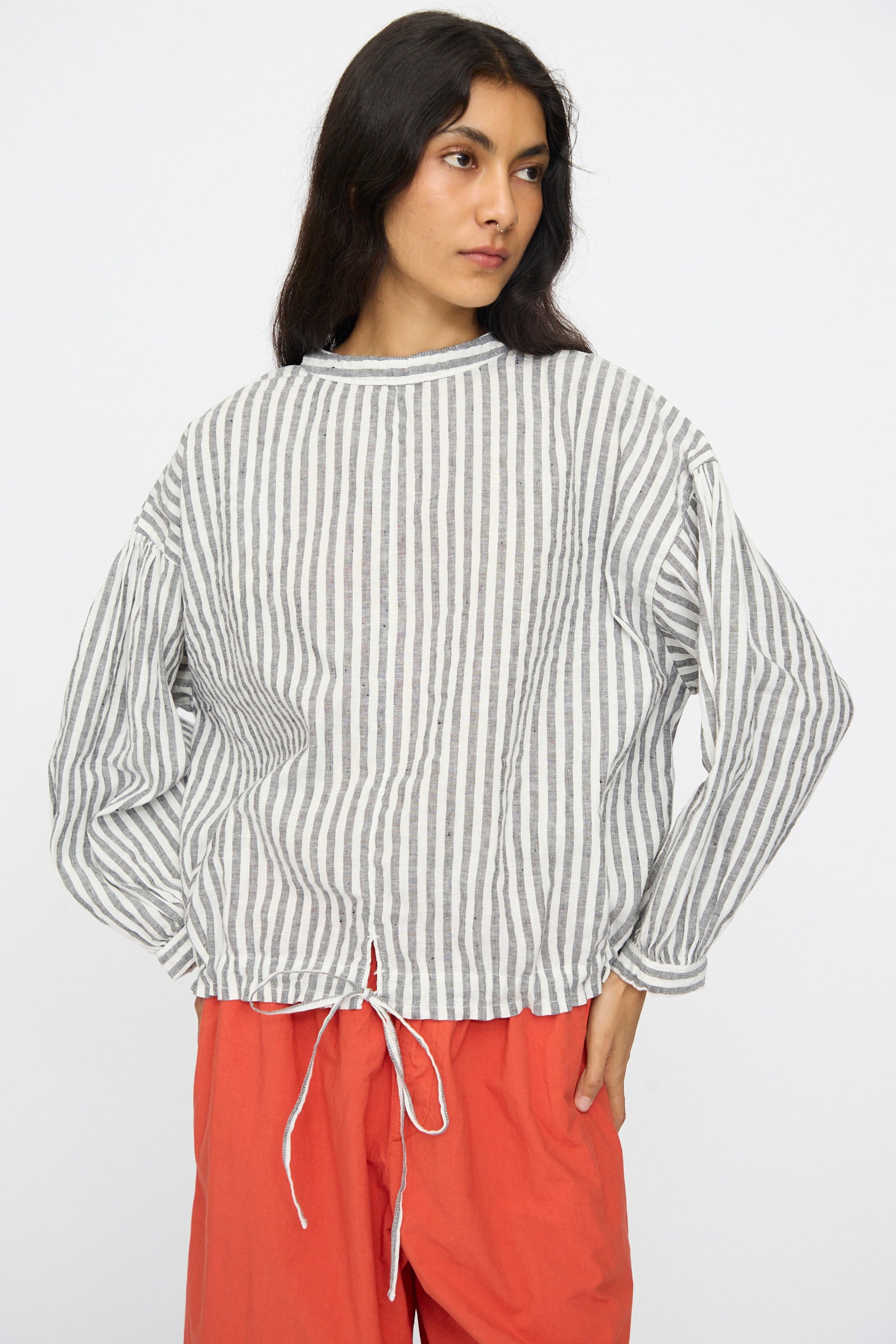 A person wears the nest Robe Upcyclelino Ribbon Two-Way Blouse in Stripe—a loose, long-sleeved gray and white striped top—paired with bright orange pants, standing against a plain white background.