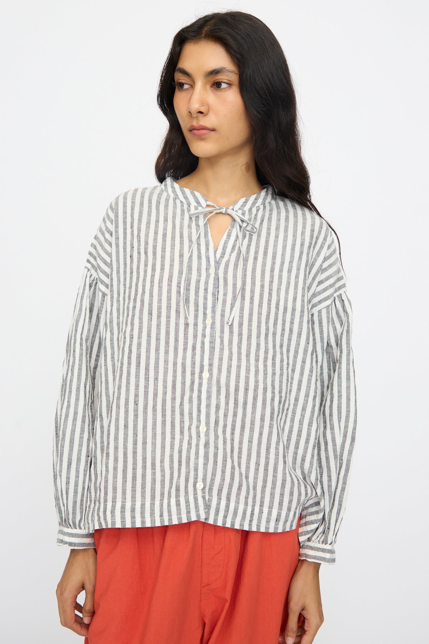 A woman models the nest Robe Upcyclelino Ribbon Two-Way Blouse in Stripe, a loose-fitting linen-cotton blend with gray and white vertical stripes, paired with rust pants against a plain white background.