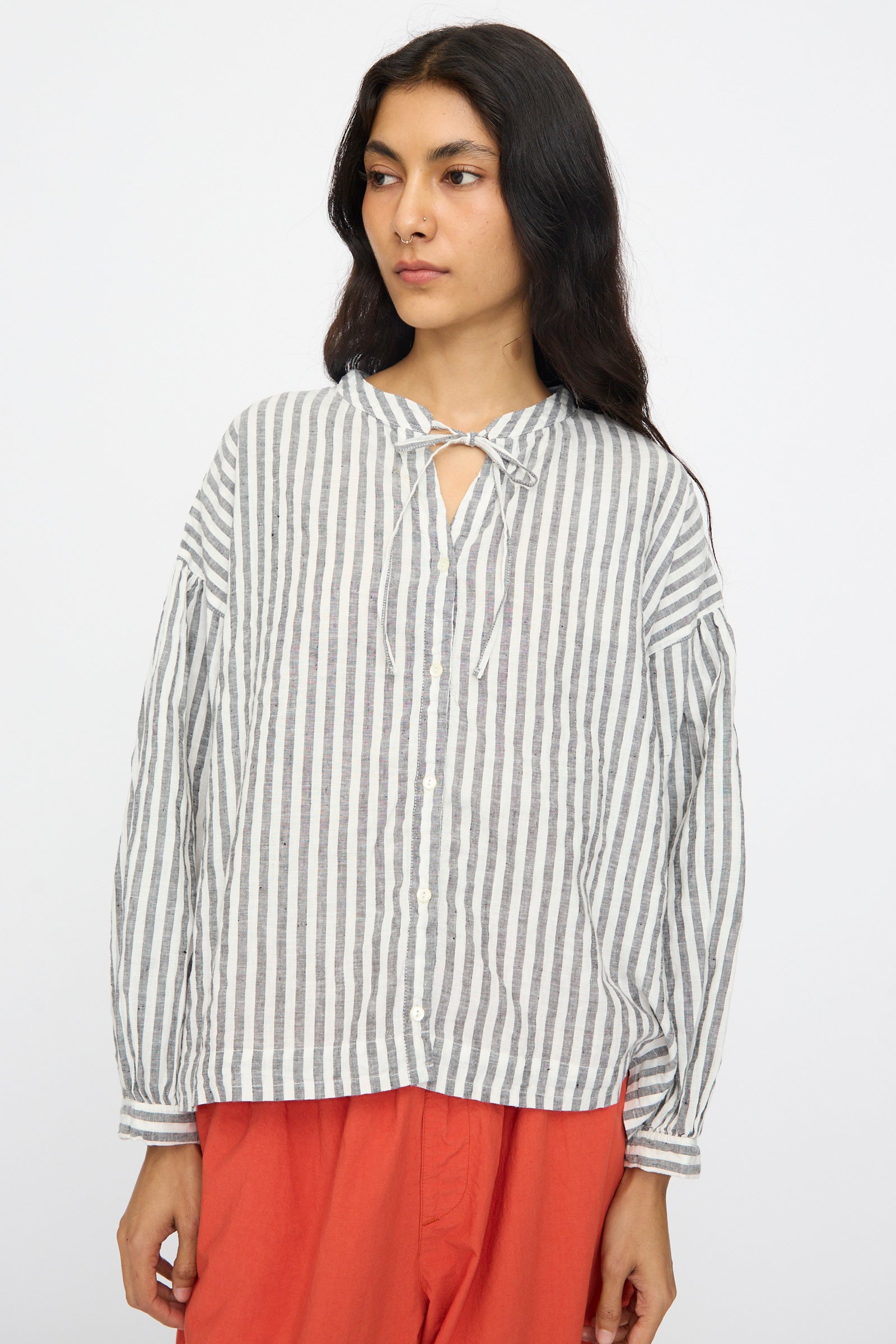 A woman models the nest Robe Upcyclelino Ribbon Two-Way Blouse in Stripe, a loose-fitting linen-cotton blend with gray and white vertical stripes, paired with rust pants against a plain white background.