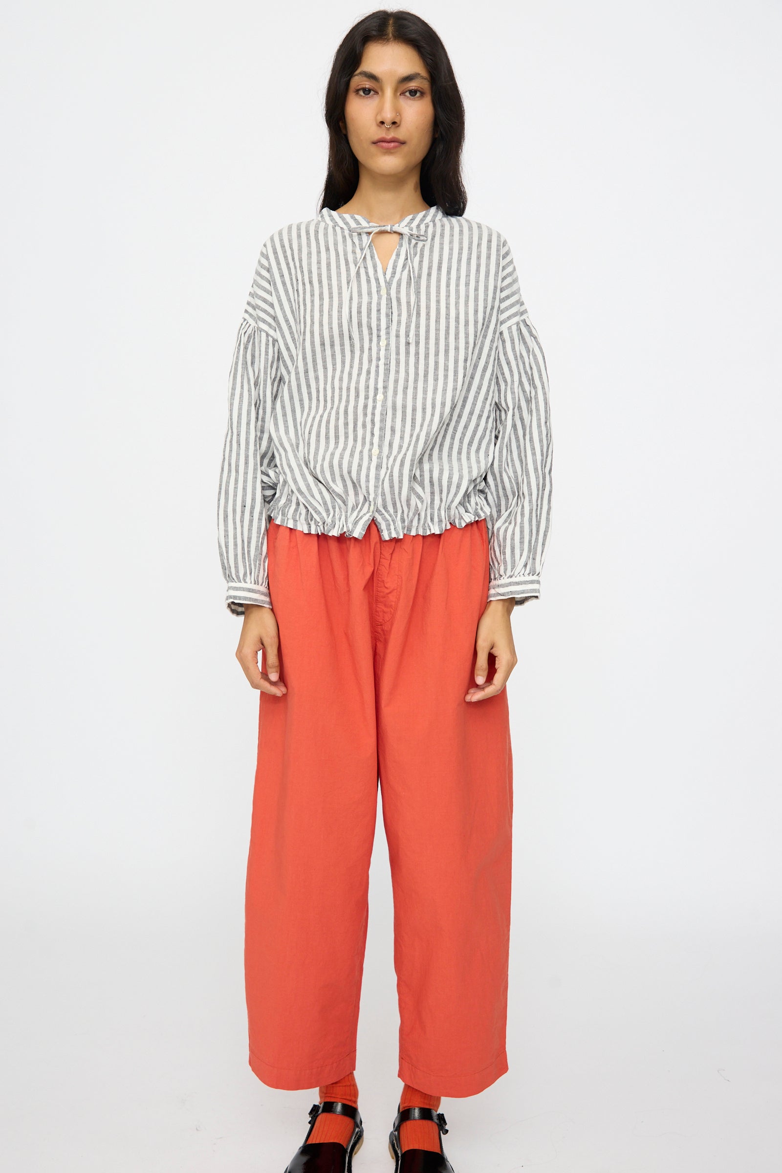 Person standing against a plain backdrop, wearing the nest Robe Upcyclelino Ribbon Two-Way Blouse in Stripe with a tie collar and adjustable hem, styled with loose red-orange pants and black sandals.