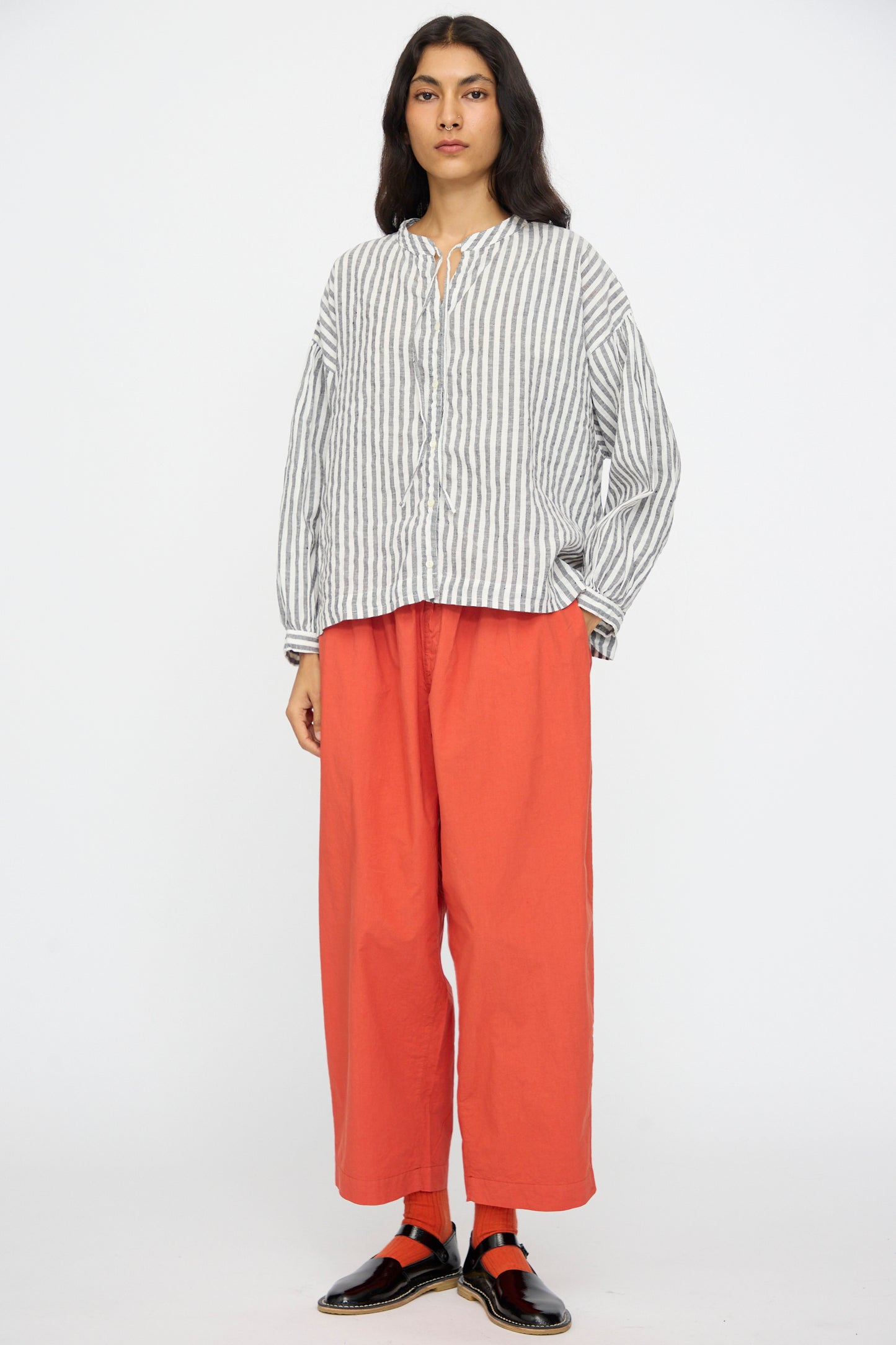 A person stands against a plain background wearing the nest Robe Upcyclelino Ribbon Two-Way Blouse in Stripe, paired with loose orange pants and black shoes with red socks.