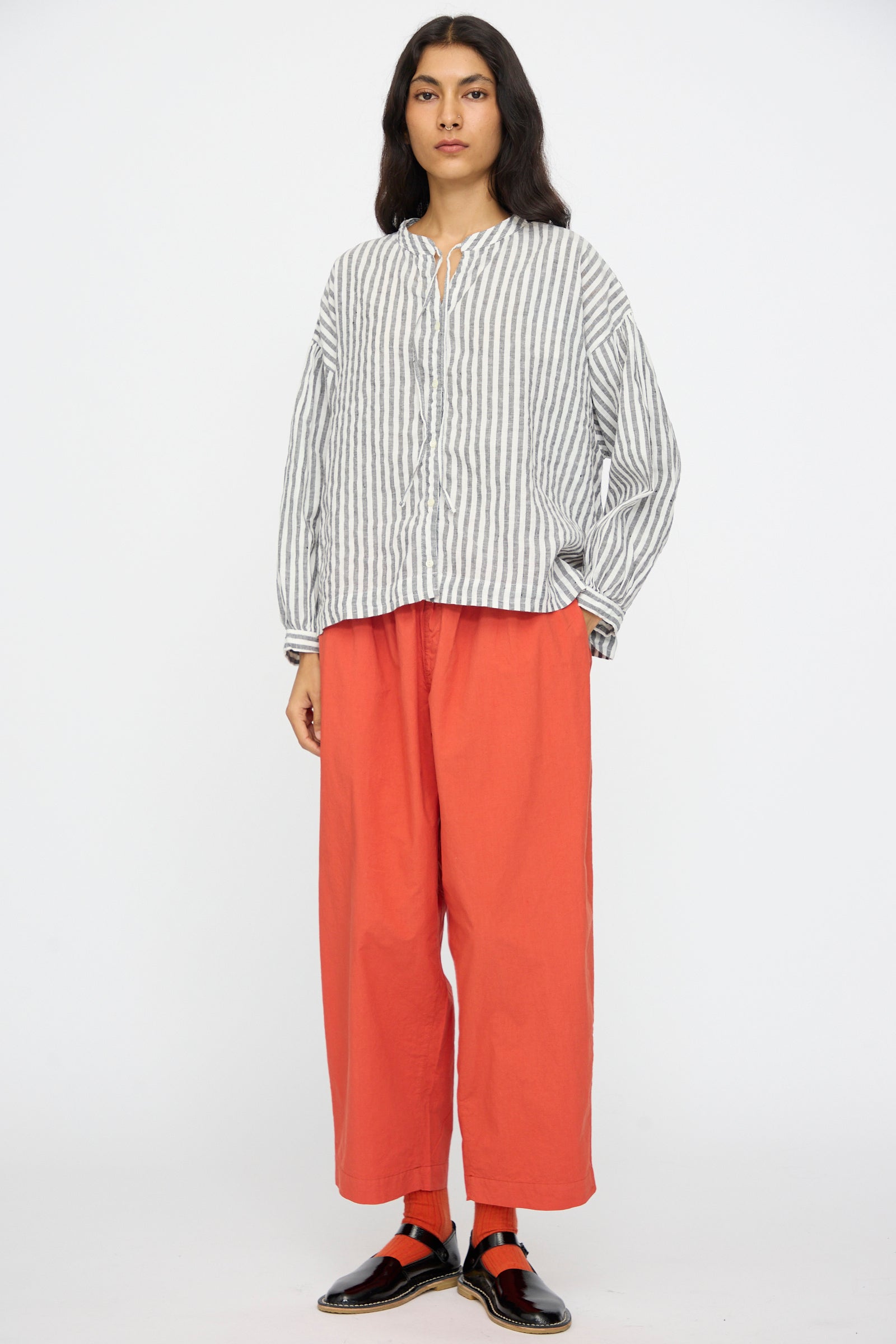 A person stands against a plain background wearing the nest Robe Upcyclelino Ribbon Two-Way Blouse in Stripe, paired with loose orange pants and black shoes with red socks.
