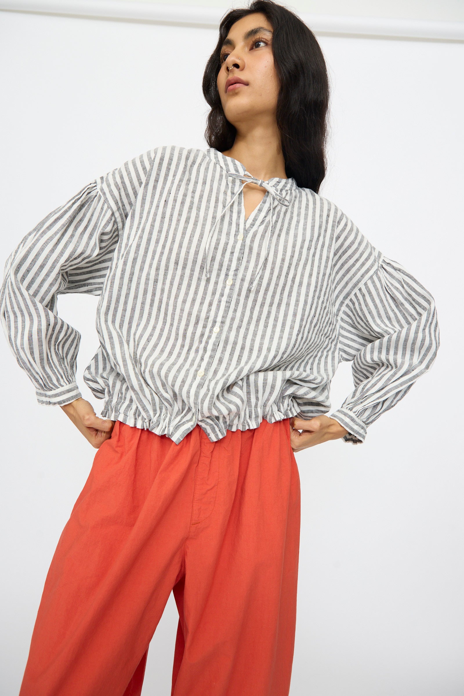 Model wears the nest Robe Upcyclelino Ribbon Two-Way Blouse in Stripe—a loose gray and white striped top with a tie neckline and adjustable hem—paired with bright red-orange wide-leg pants, standing against a plain white background.