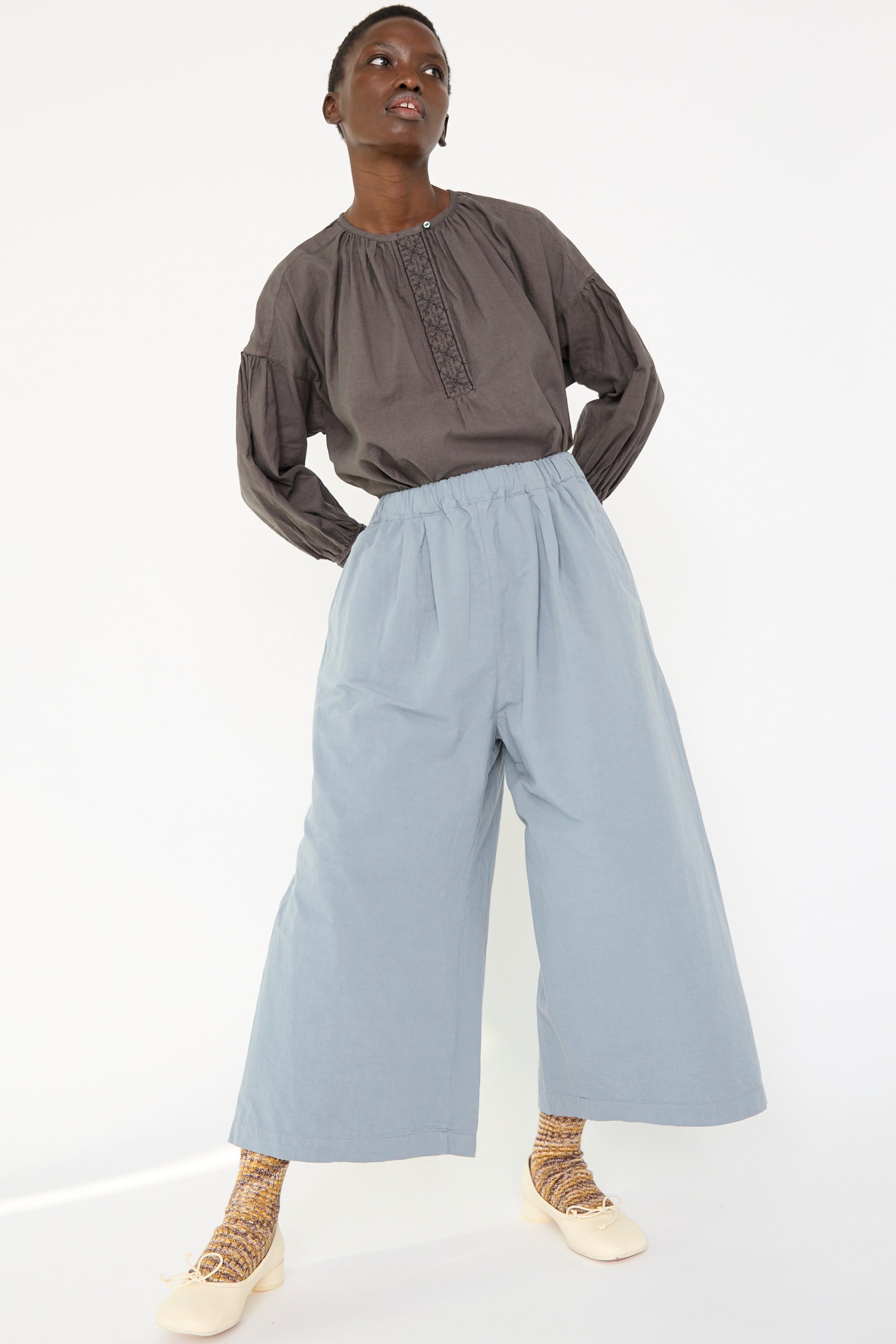 A woman wears a loose-fitting gray blouse paired with nest Robe's Water Repellent Cotton Linen Omi-Zarahi Wide Pant in Blue, along with patterned socks and beige shoes, set against a plain background.